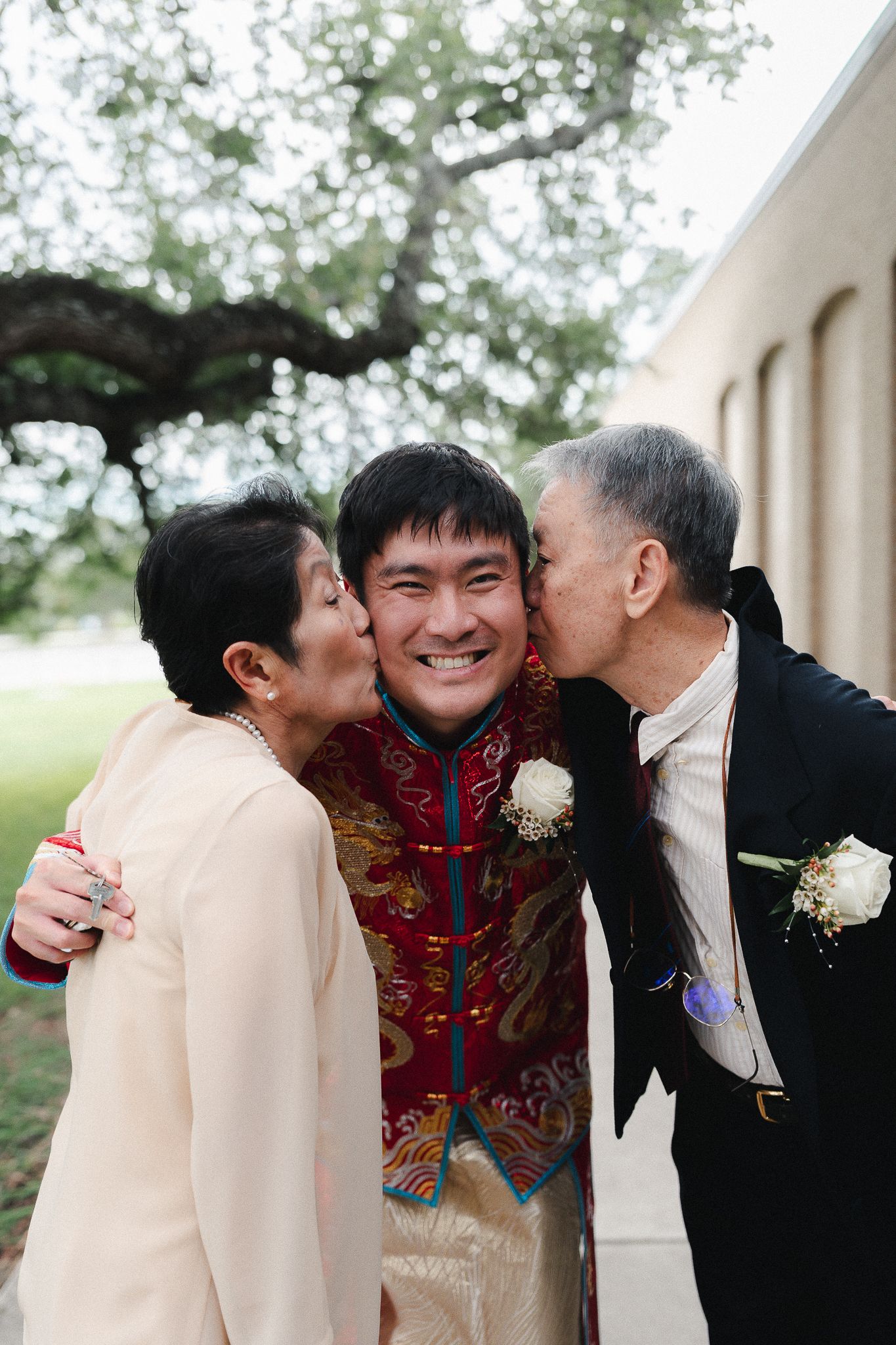 Groom in traditional Chinese wedding attire receives kisses on cheeks from proud parents outdoors.