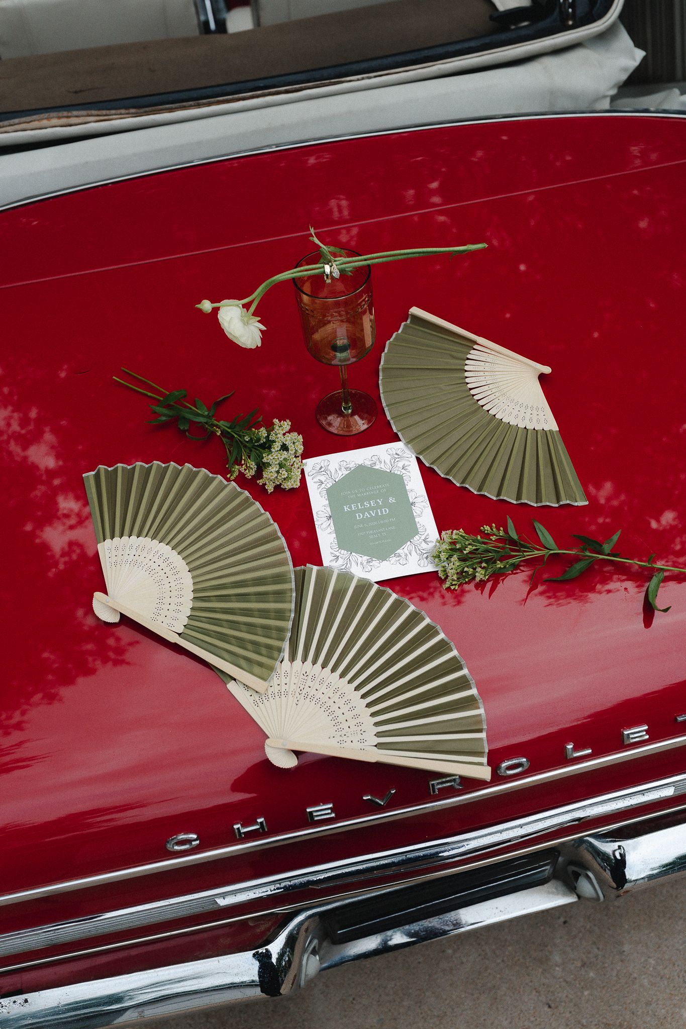 Olive green hand fans and a red goblet displayed on a vintage red Chevrolet trunk with floral accents and wedding card.