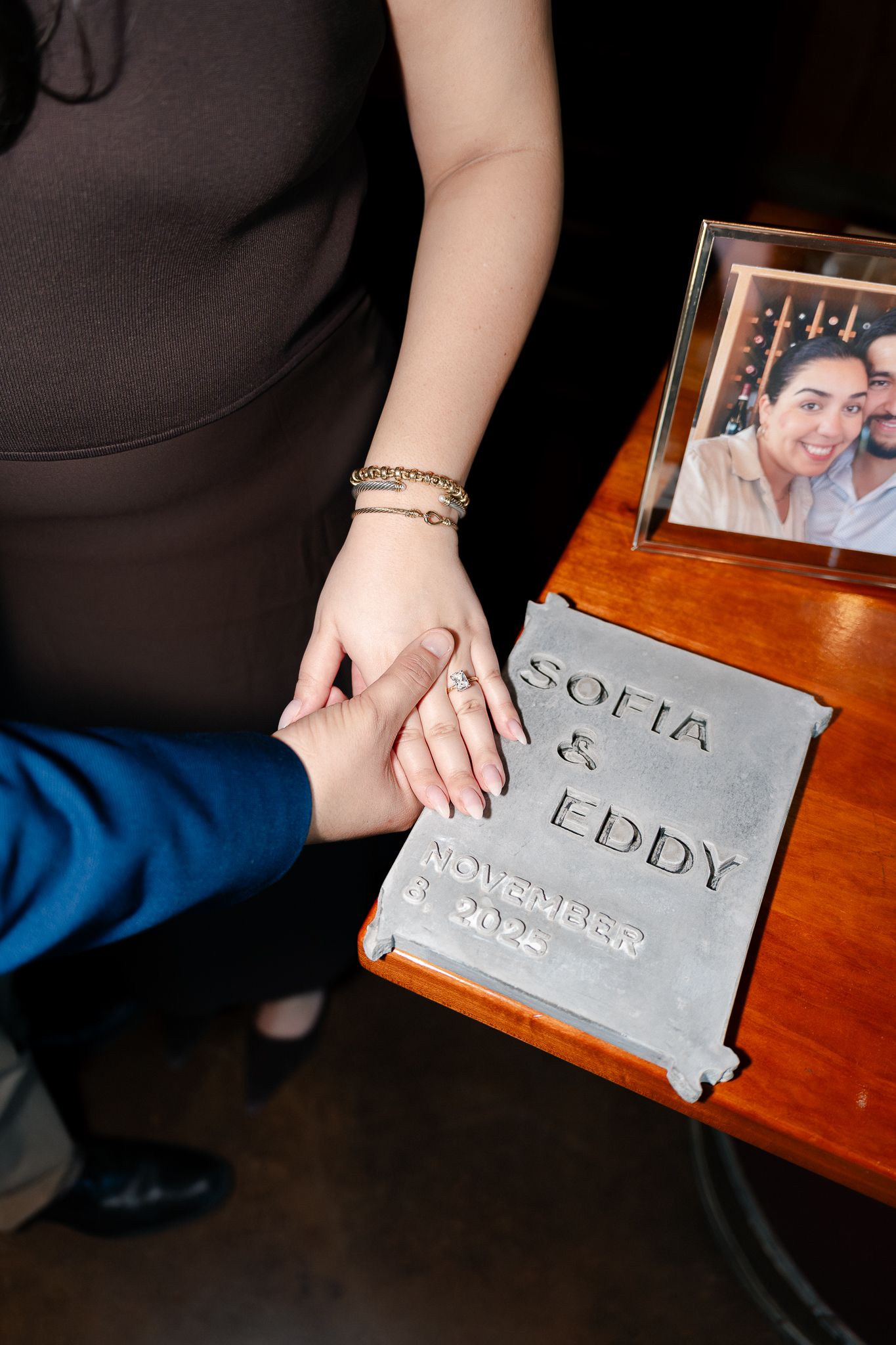 A hand traces letters written in dust on a wooden table surface next to framed photographs.