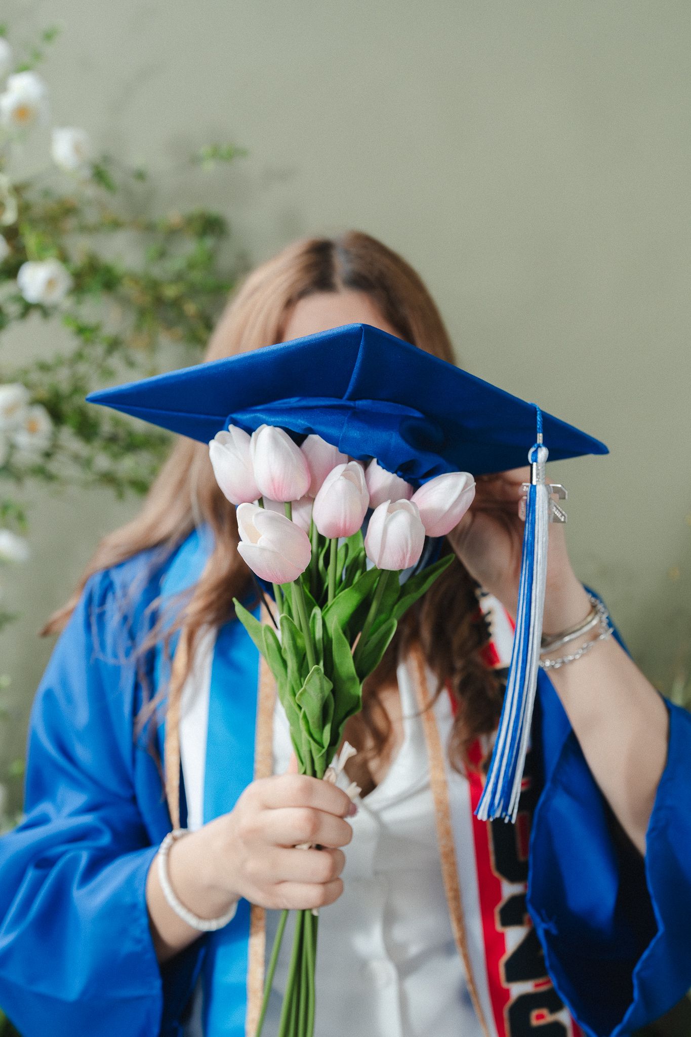 Graduate in blue cap and gown holds pink tulip bouquet covering face, celebrating graduation day.