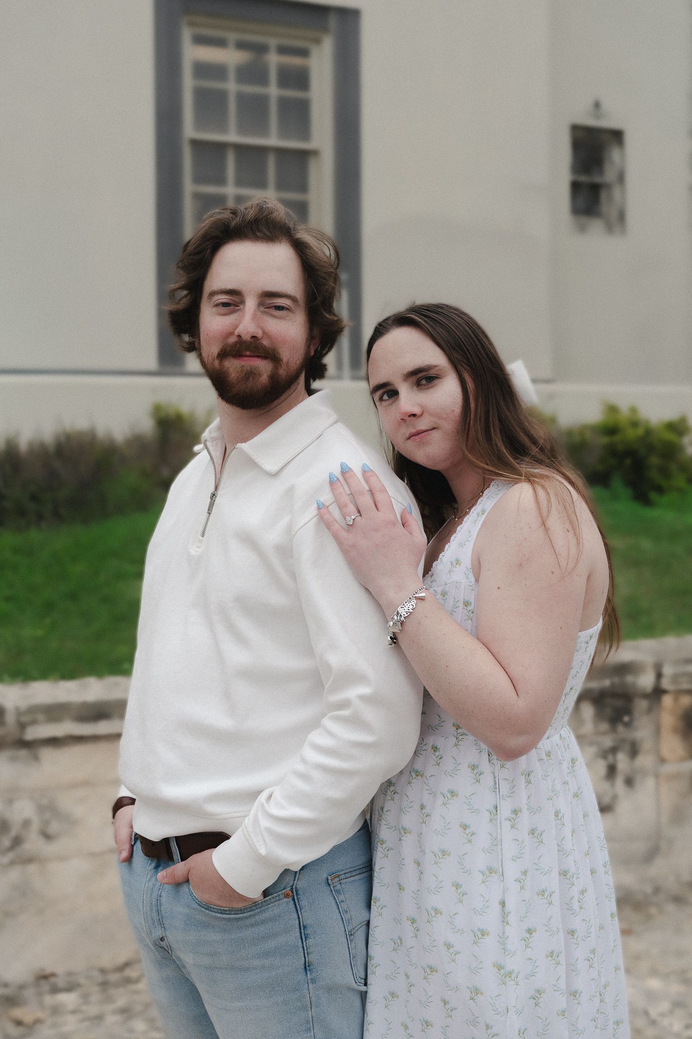 Couple posing outdoors, man in white shirt and jeans, woman in floral dress leaning on his shoulder.