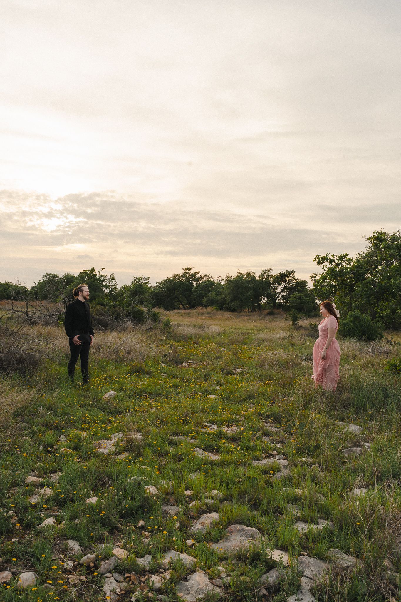 A man in black and woman in pink dress stand apart in a scenic green field at dusk.