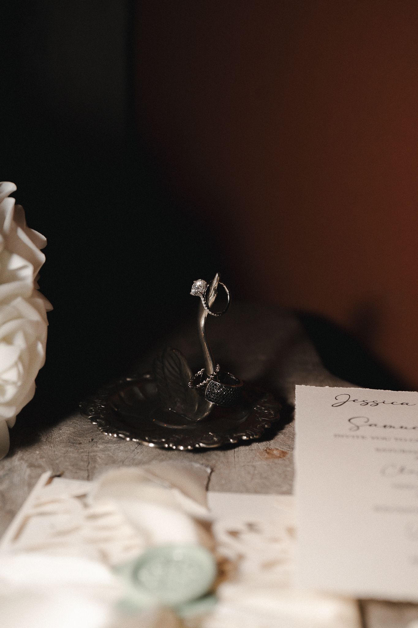 Diamond engagement ring on a silver leaf-shaped ring holder beside a wedding invitation, wax seal and white rose on a rustic table.