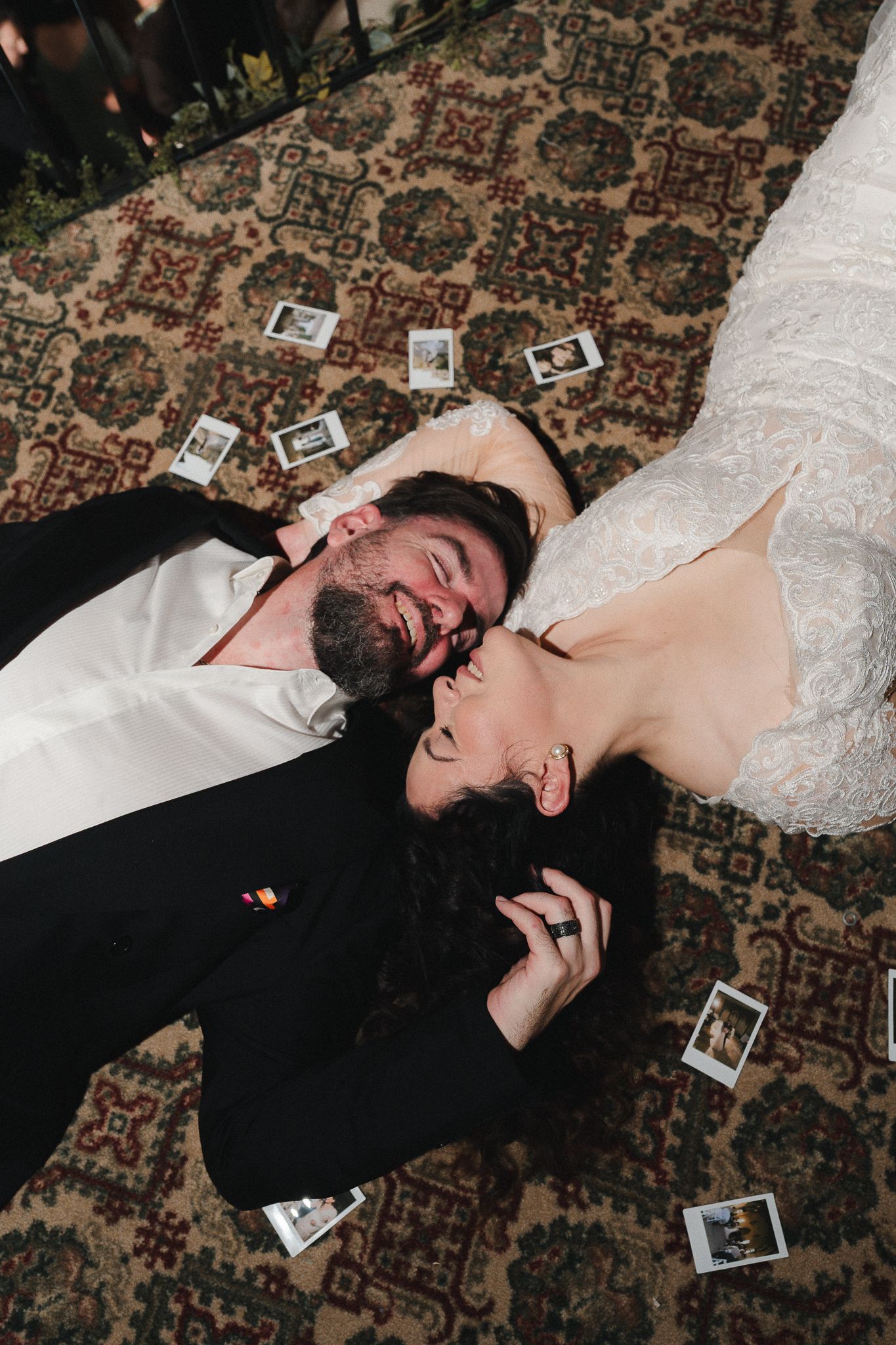 Smiling bride in a white lace gown and groom in a black suit lie head to head on a patterned rug scattered with instant photos.
