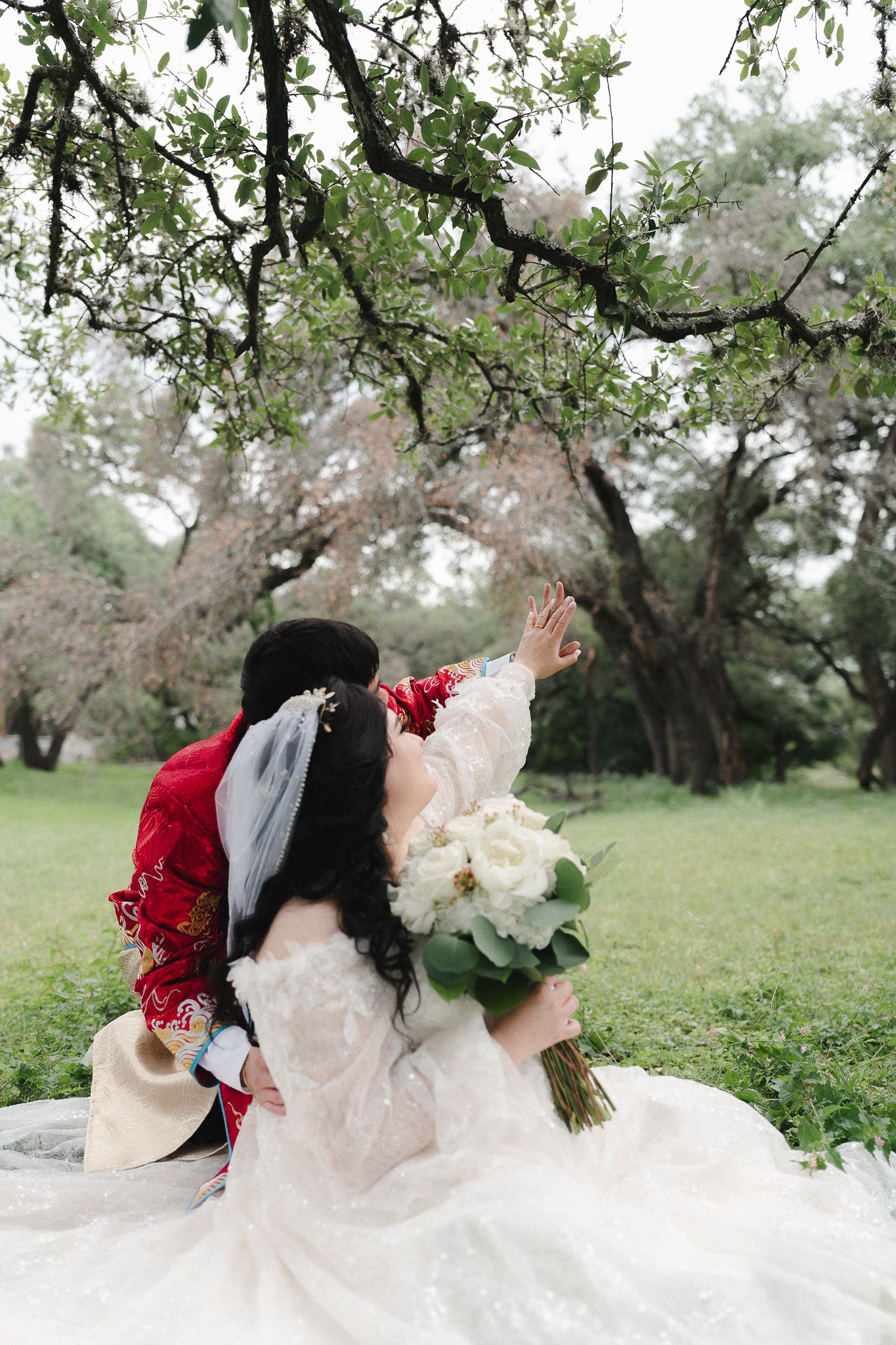 Bride in white lace gown holding bouquet sits with groom in red traditional attire under flowering tree in garden.