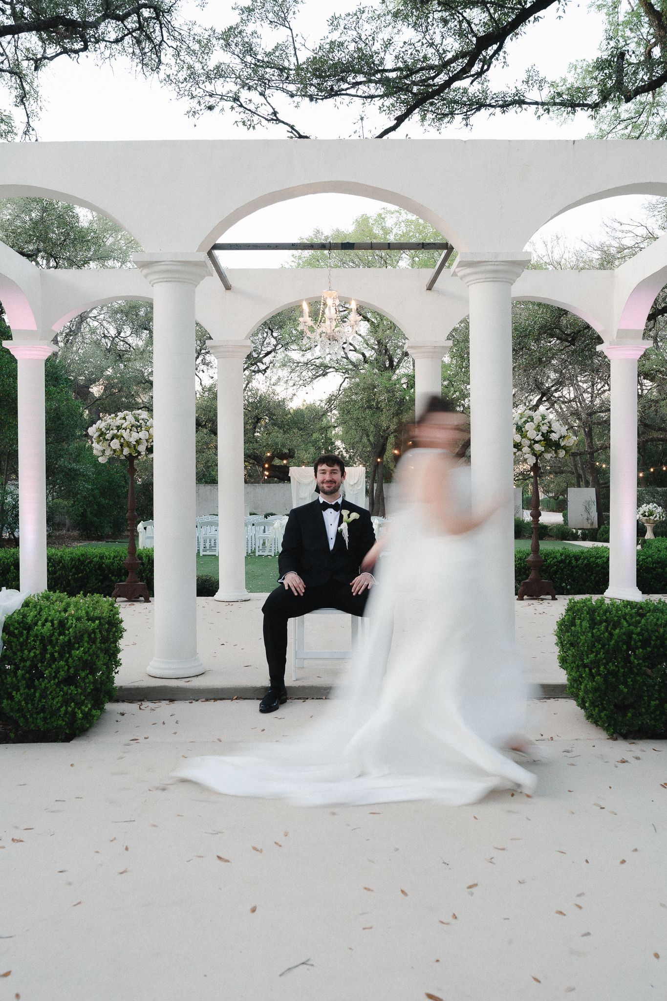 Groom in tuxedo sits under white arched colonnade as bride twirls in flowing white gown at elegant outdoor wedding venue.