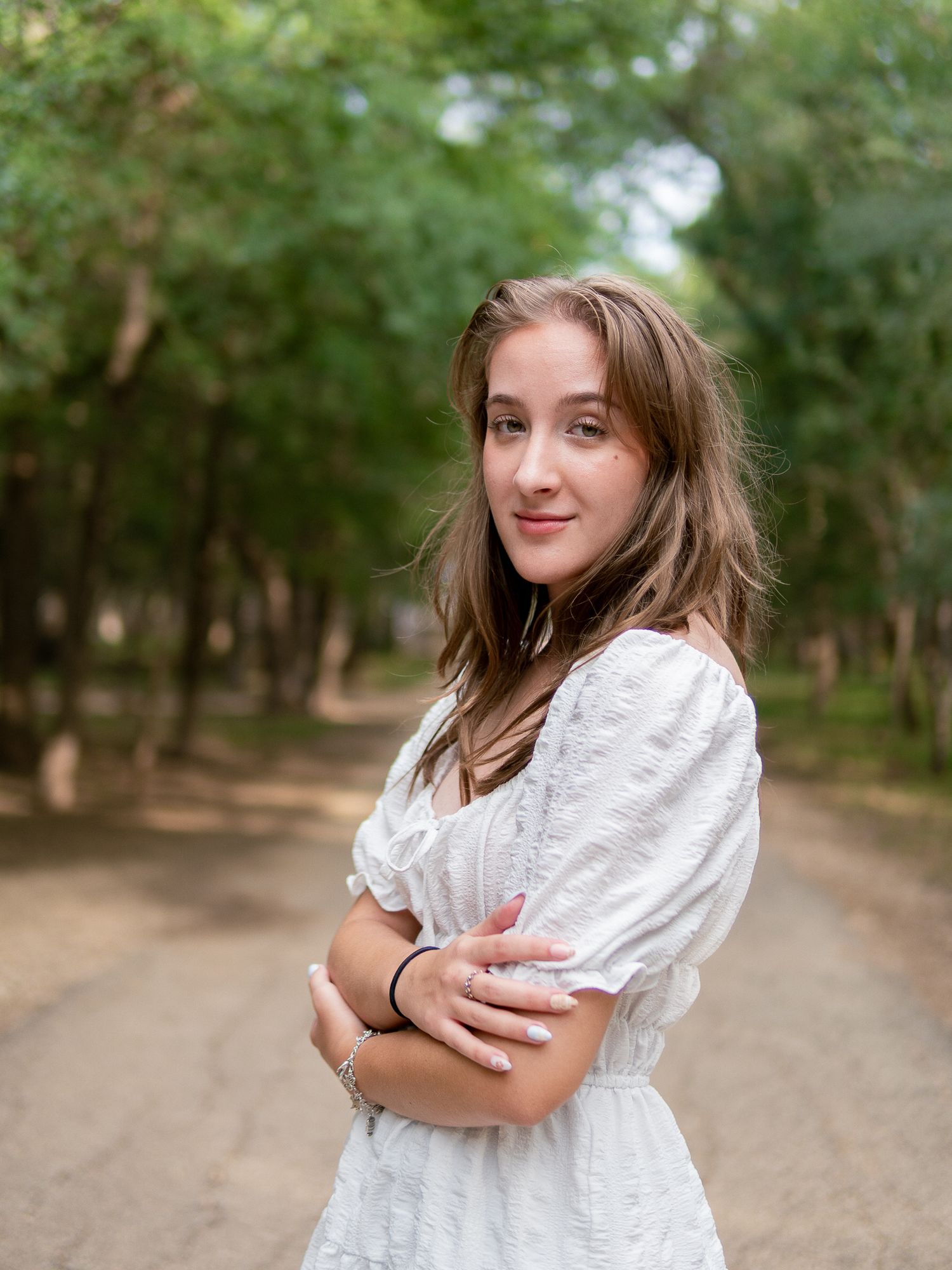 A person in a white lace dress stands on a dirt path surrounded by green trees in a forest setting.