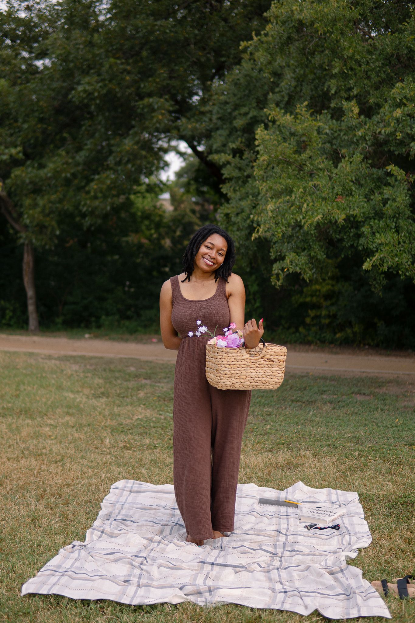 A person in a brown outfit sits on a picnic blanket in a park setting with trees in the background.