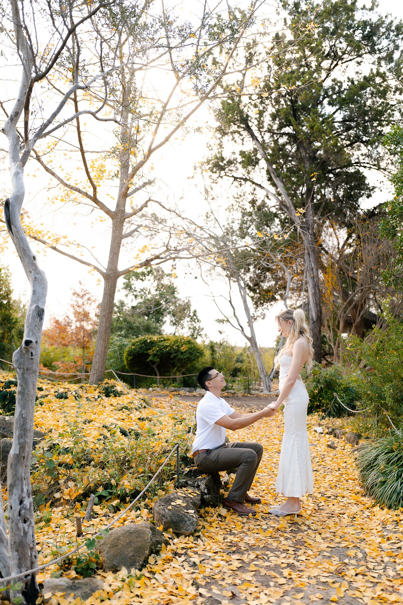 A couple shares a romantic moment surrounded by fallen autumn leaves in a scenic outdoor setting.