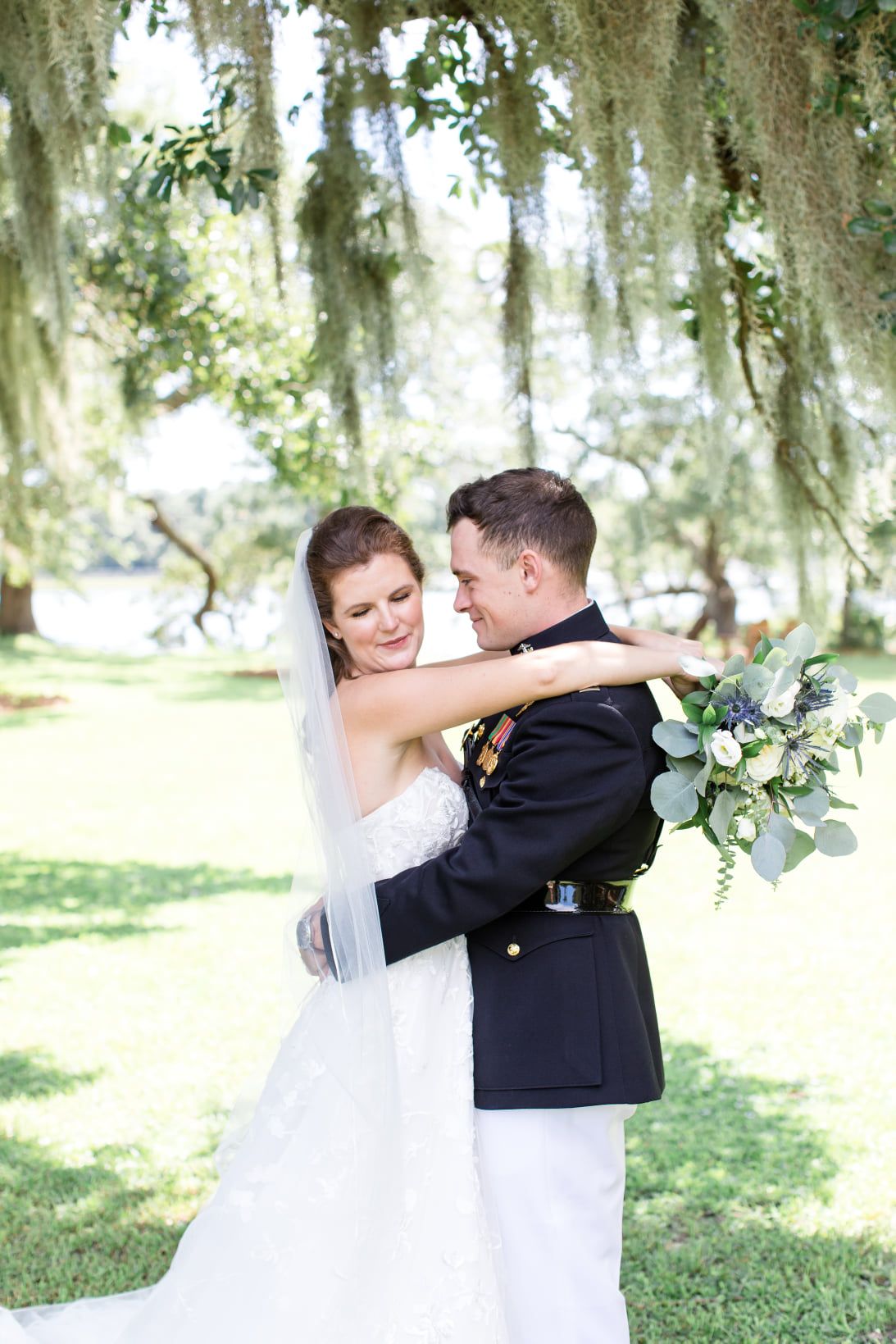 A couple embraces under Spanish moss at an elegant outdoor wedding featuring a white dress and military dress uniform.