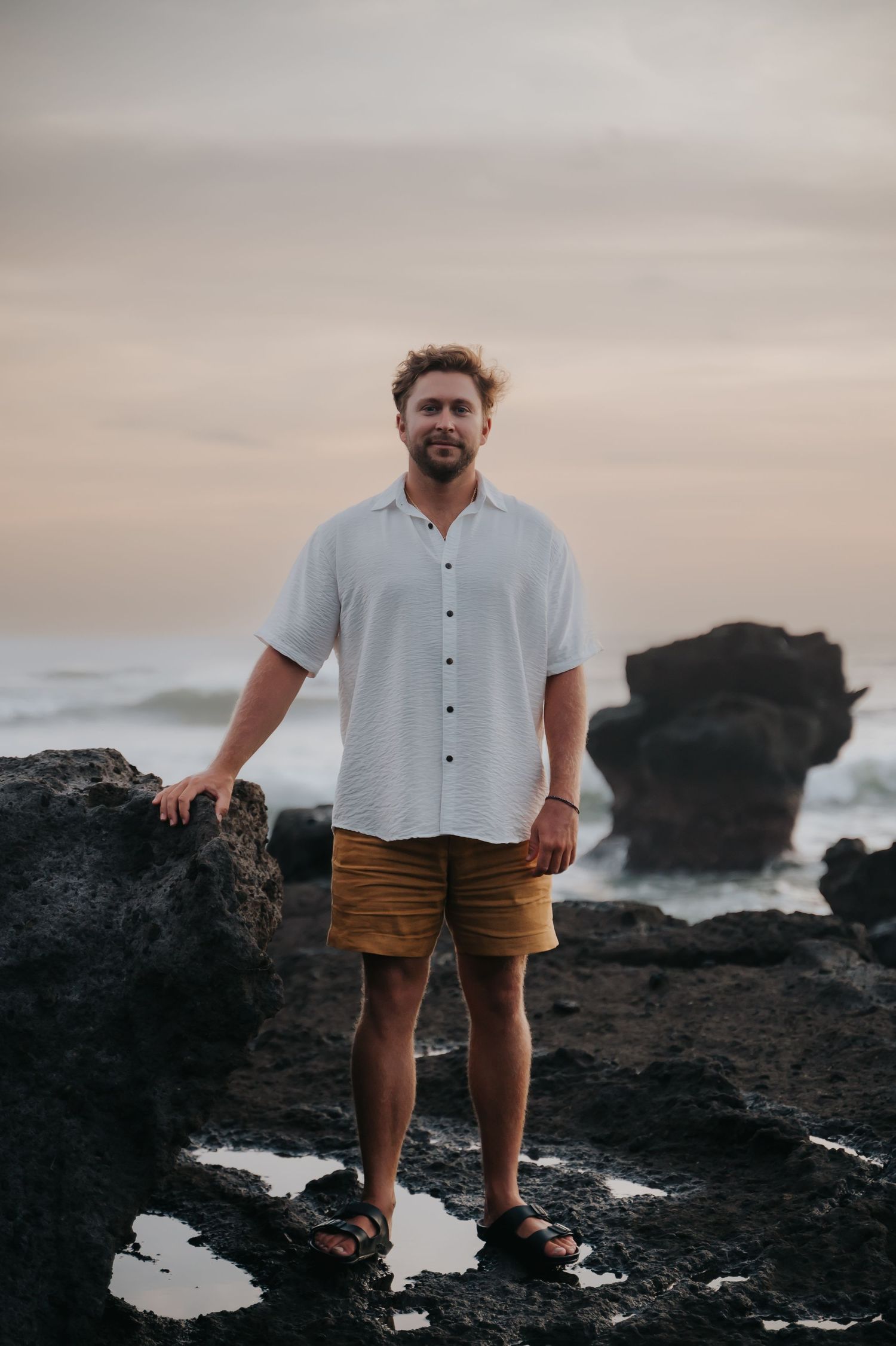 Someone in a white button-down shirt and brown shorts stands on rocky coastal terrain at sunset with ocean waves behind.