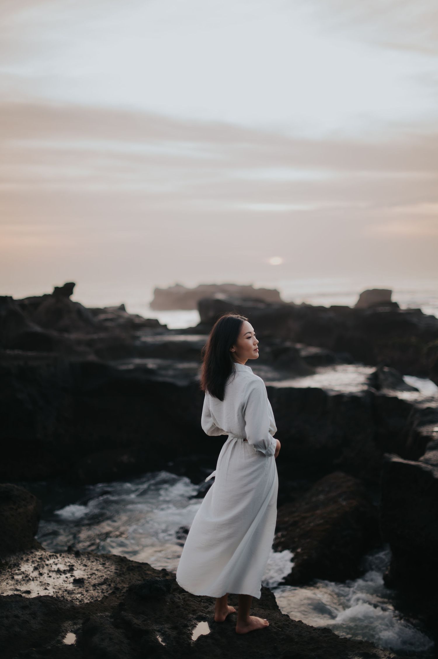 A figure in a flowing white dress dances gracefully on dark rocky coastal cliffs at sunset.