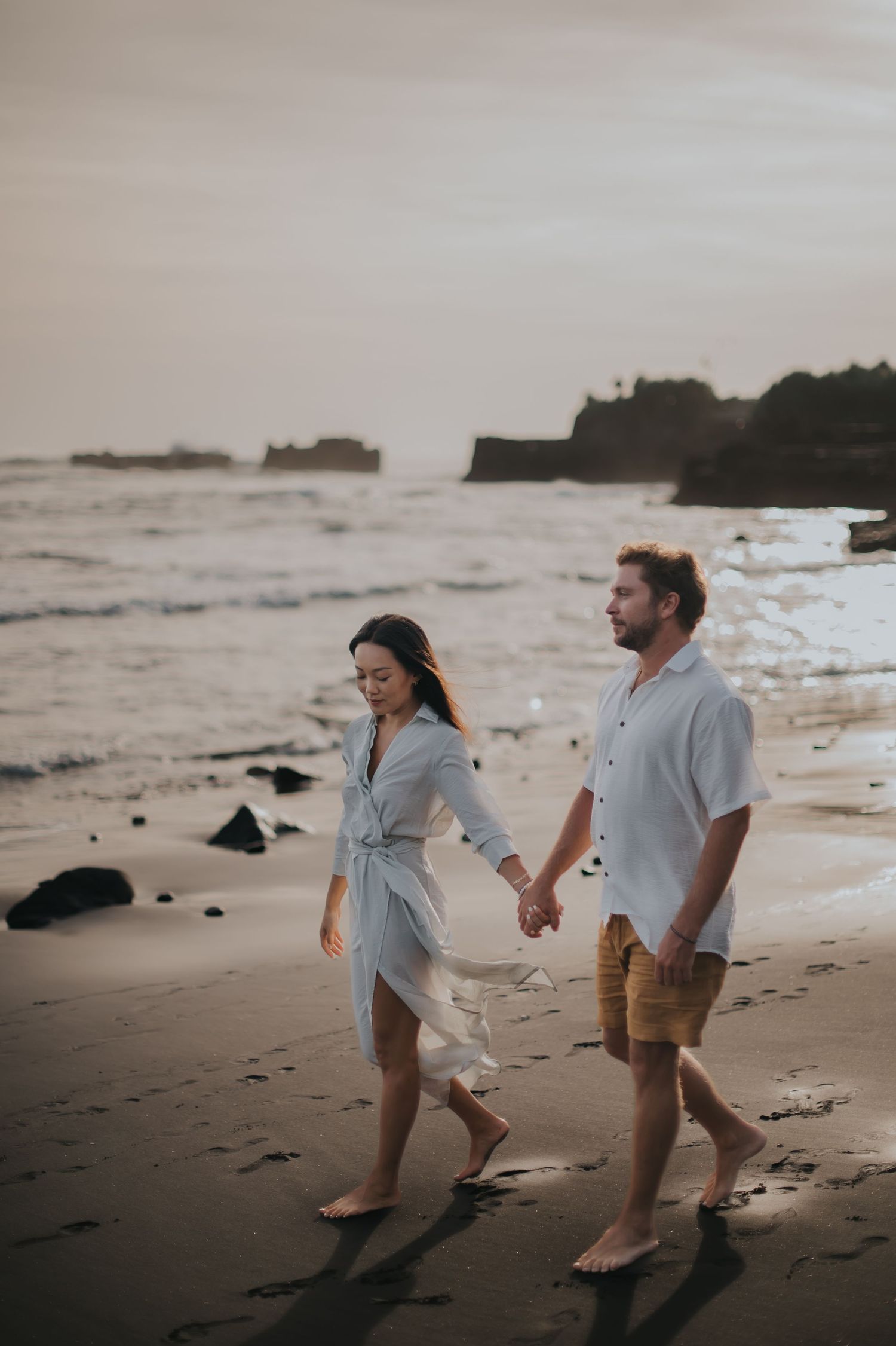 A couple walks hand in hand along a misty beach at sunset with cliffs and ocean waves in the background.
