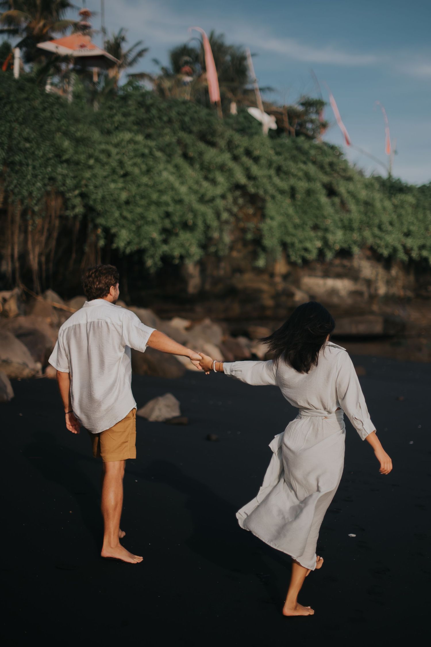 Two people dancing barefoot on a rocky beach with palm trees and buildings visible on a cliff in the background.