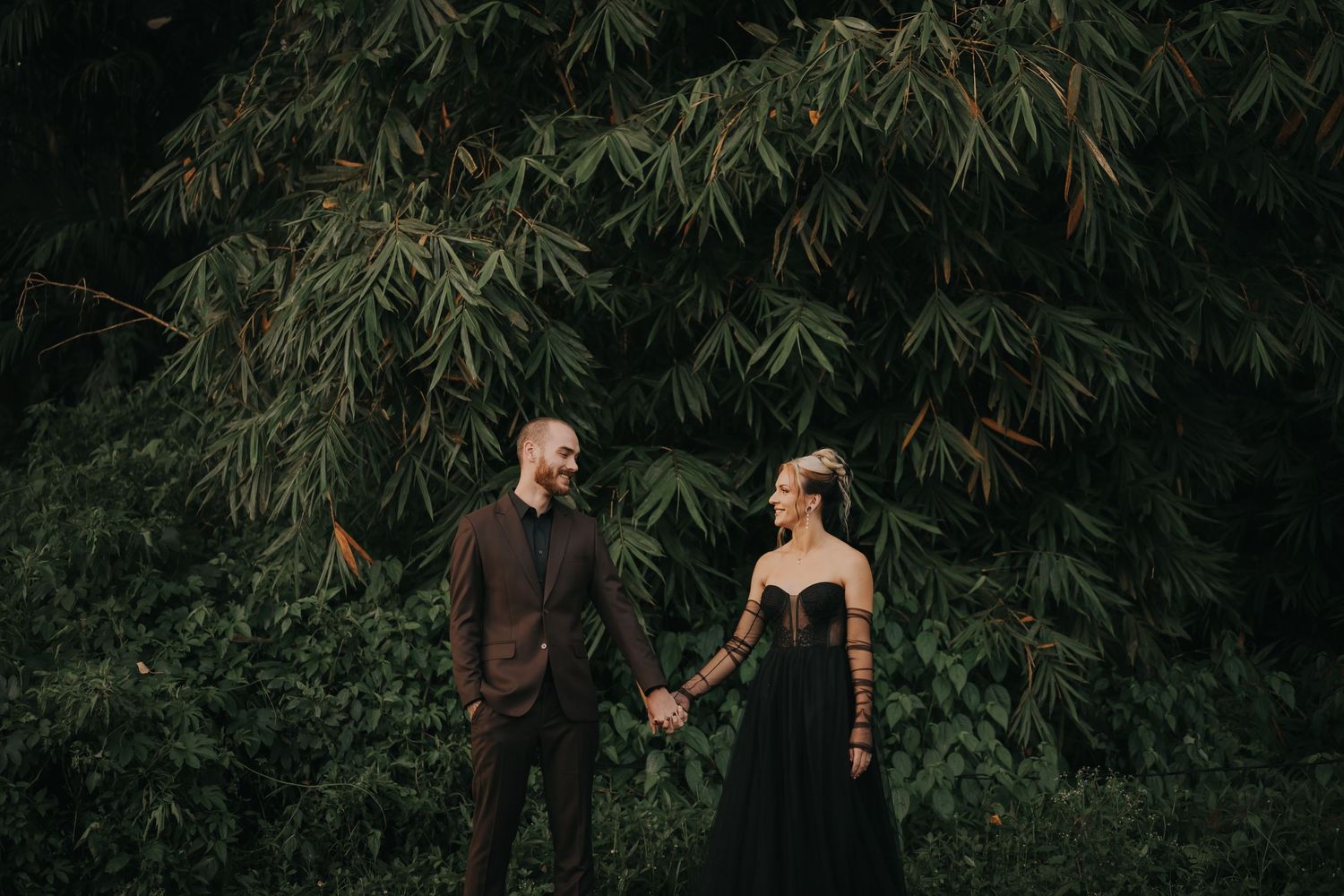 Couple in formal attire poses together in a lush garden setting with bamboo plants in the background.