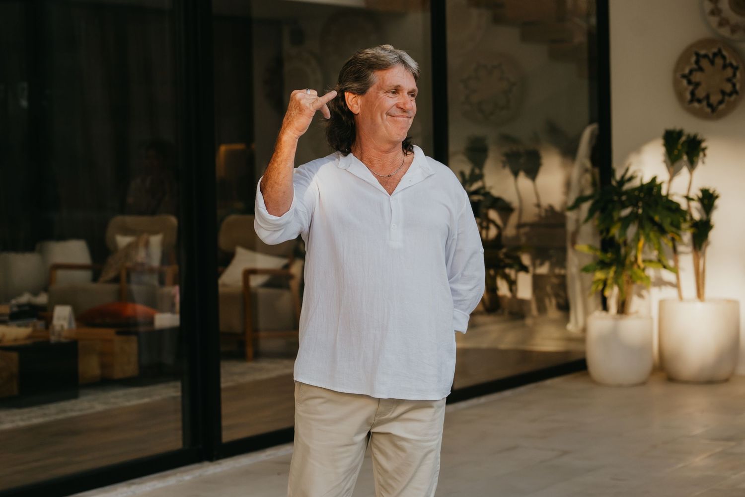 Smiling middle-aged man in white linen shirt gesturing while standing in a modern tropical villa interior.