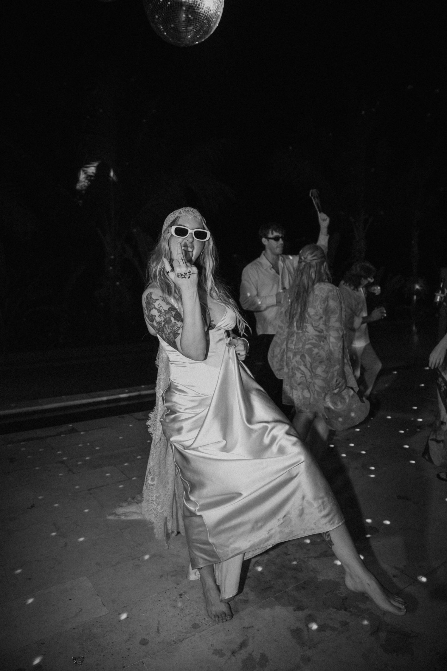 Bride in satin dress and sunglasses dances barefoot under disco ball at lively wedding reception party.