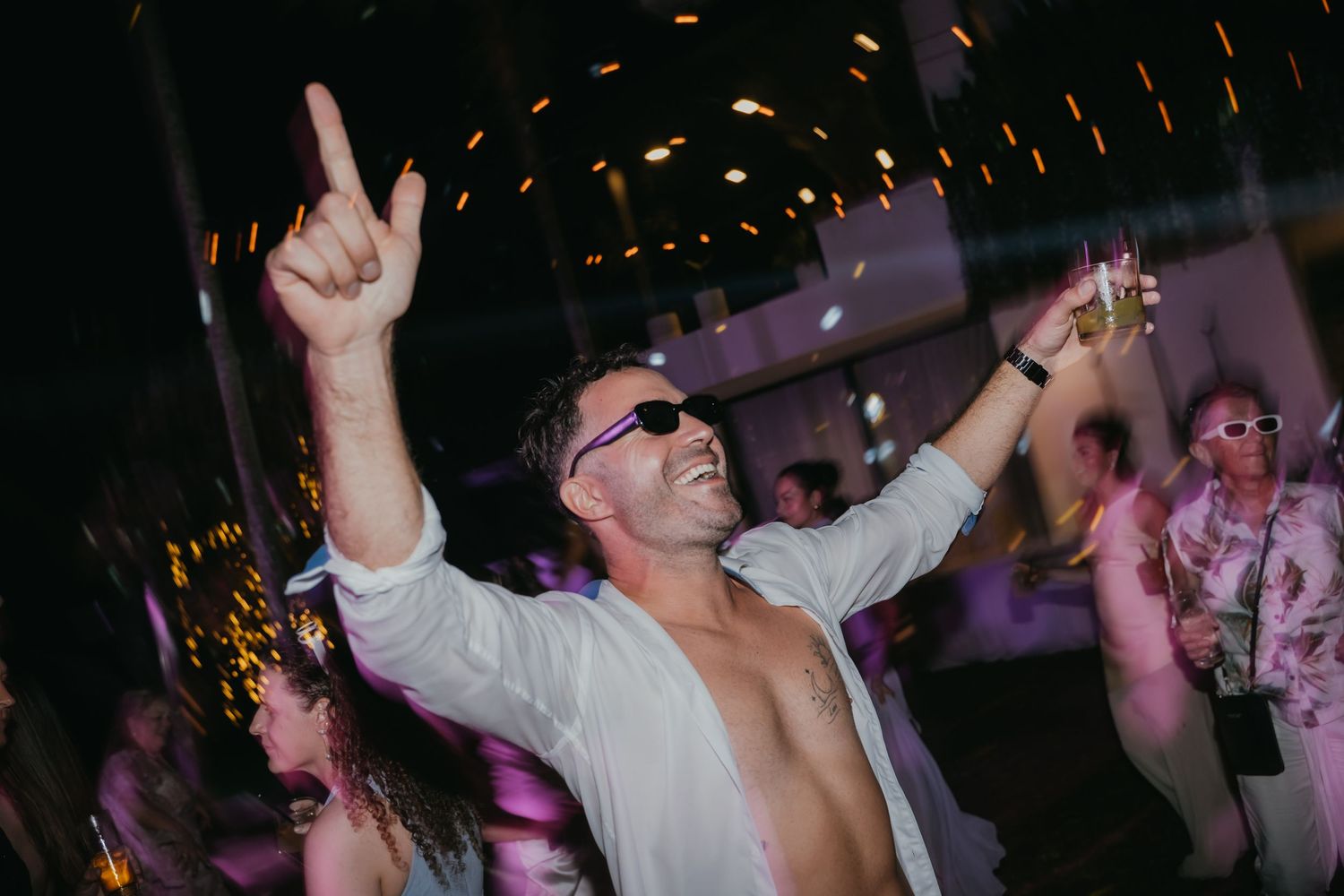 Energetic wedding guest dancing with sunglasses and raised arms at reception party.