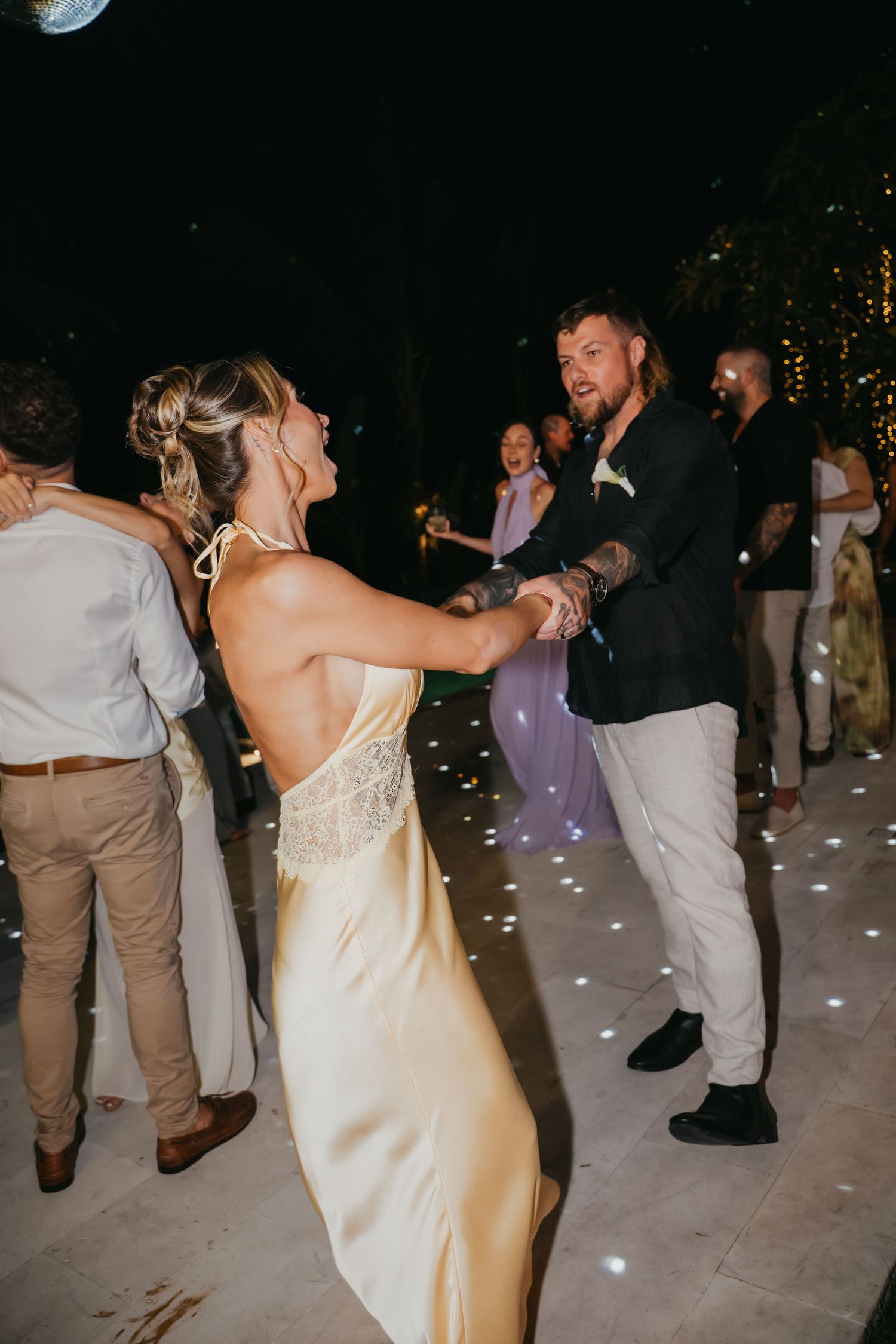 Bride in gold satin gown dances with groom on illuminated dance floor at outdoor wedding reception.