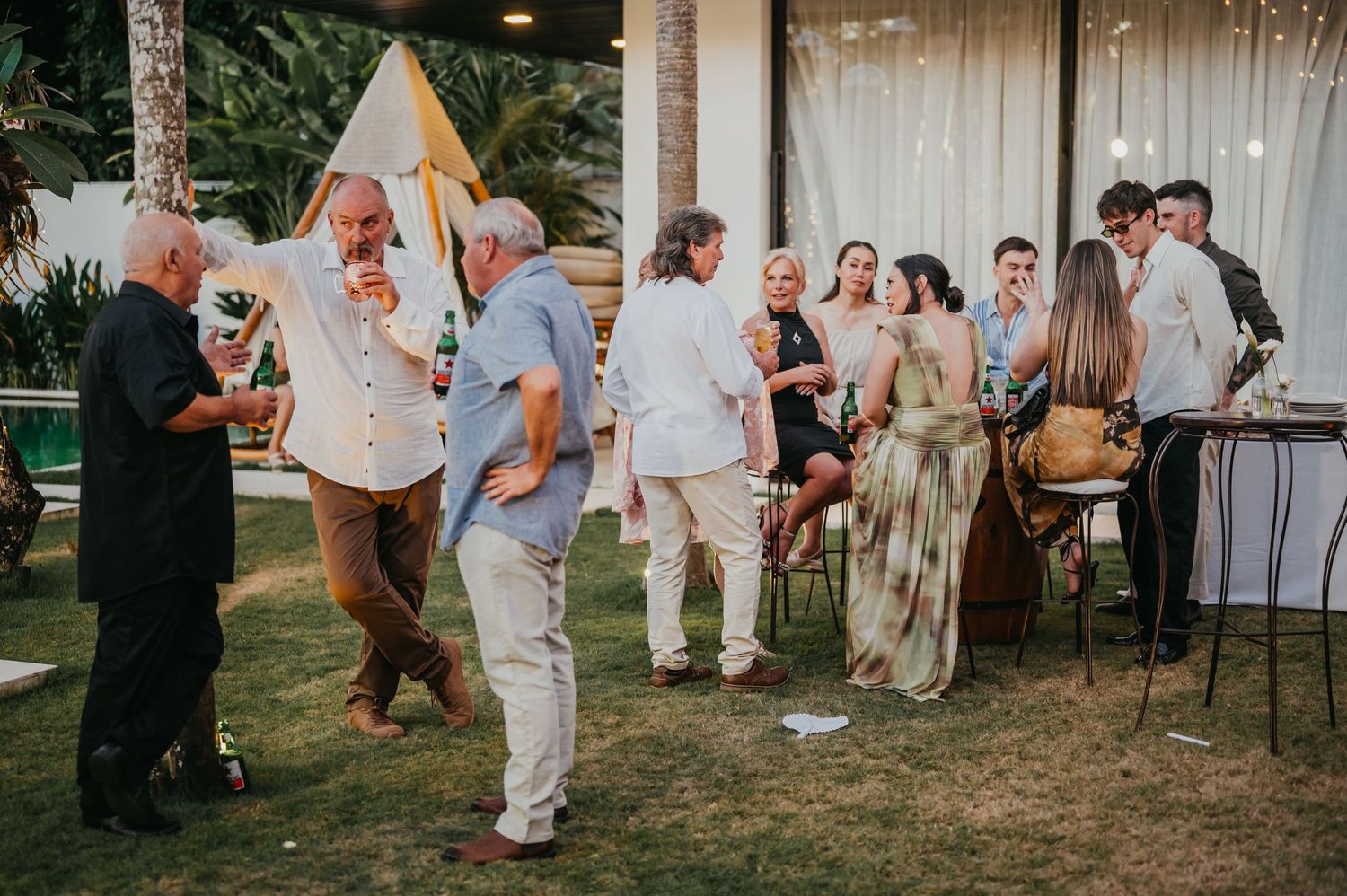Wedding guests mingle on lush green lawn during outdoor tropical reception cocktail hour.