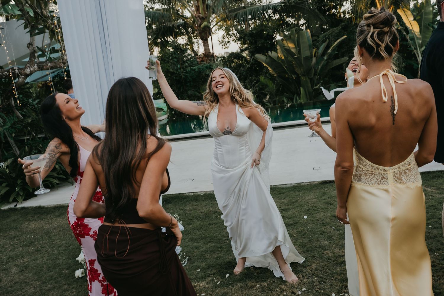 Bride in white gown laughing and dancing with bridesmaids on the lawn during outdoor wedding reception.