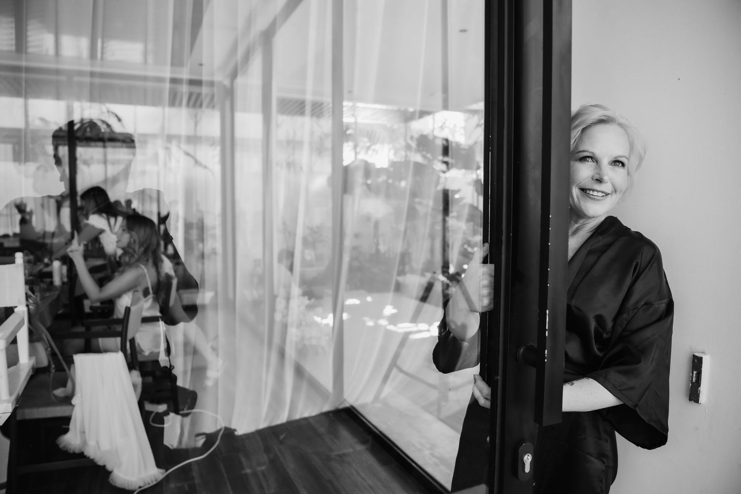 Bride in black robe smiling by glass door during wedding preparations in black and white photo.