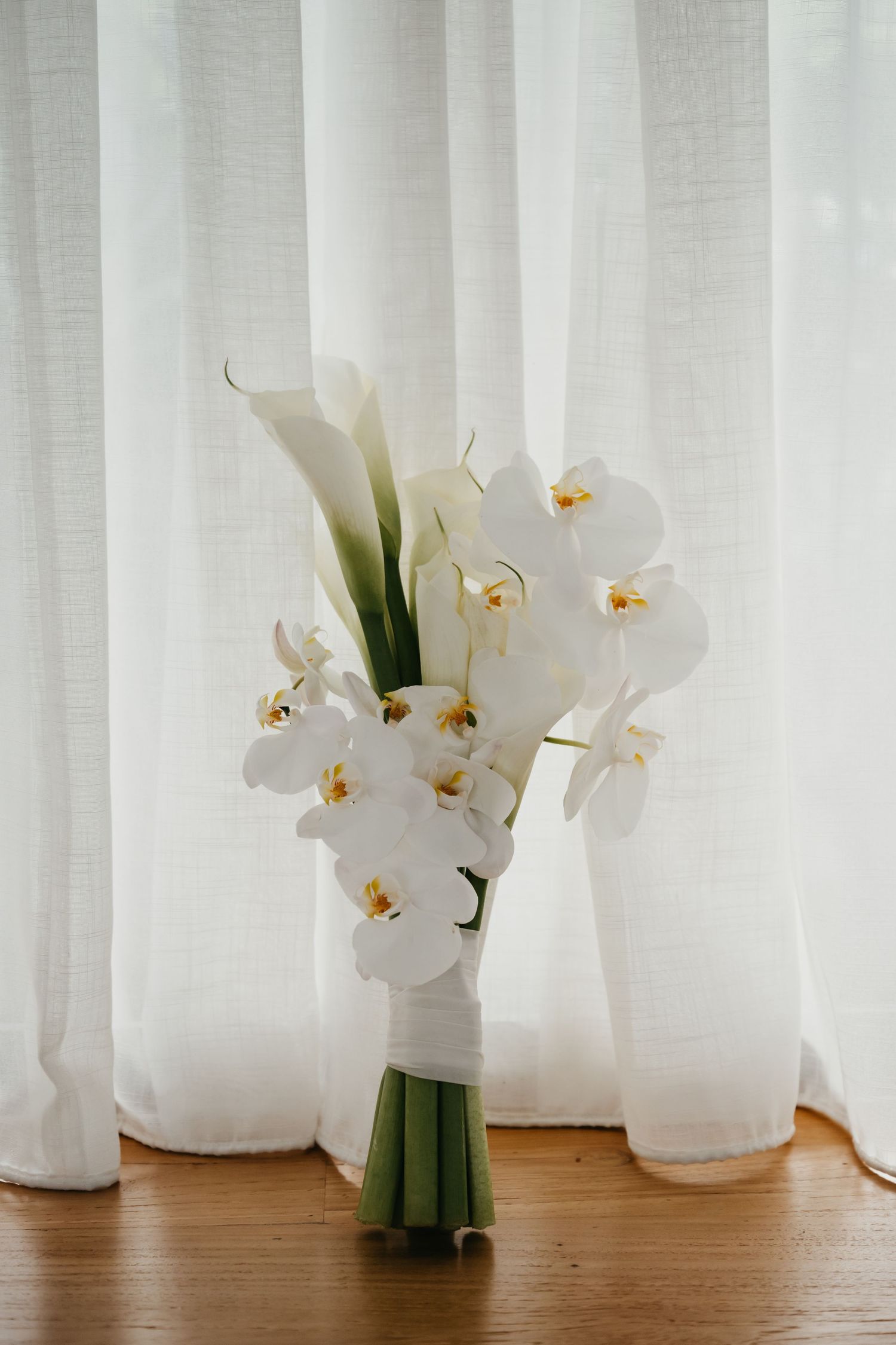 Elegant bridal bouquet of white orchids and calla lilies tied with green stems against sheer white curtains.