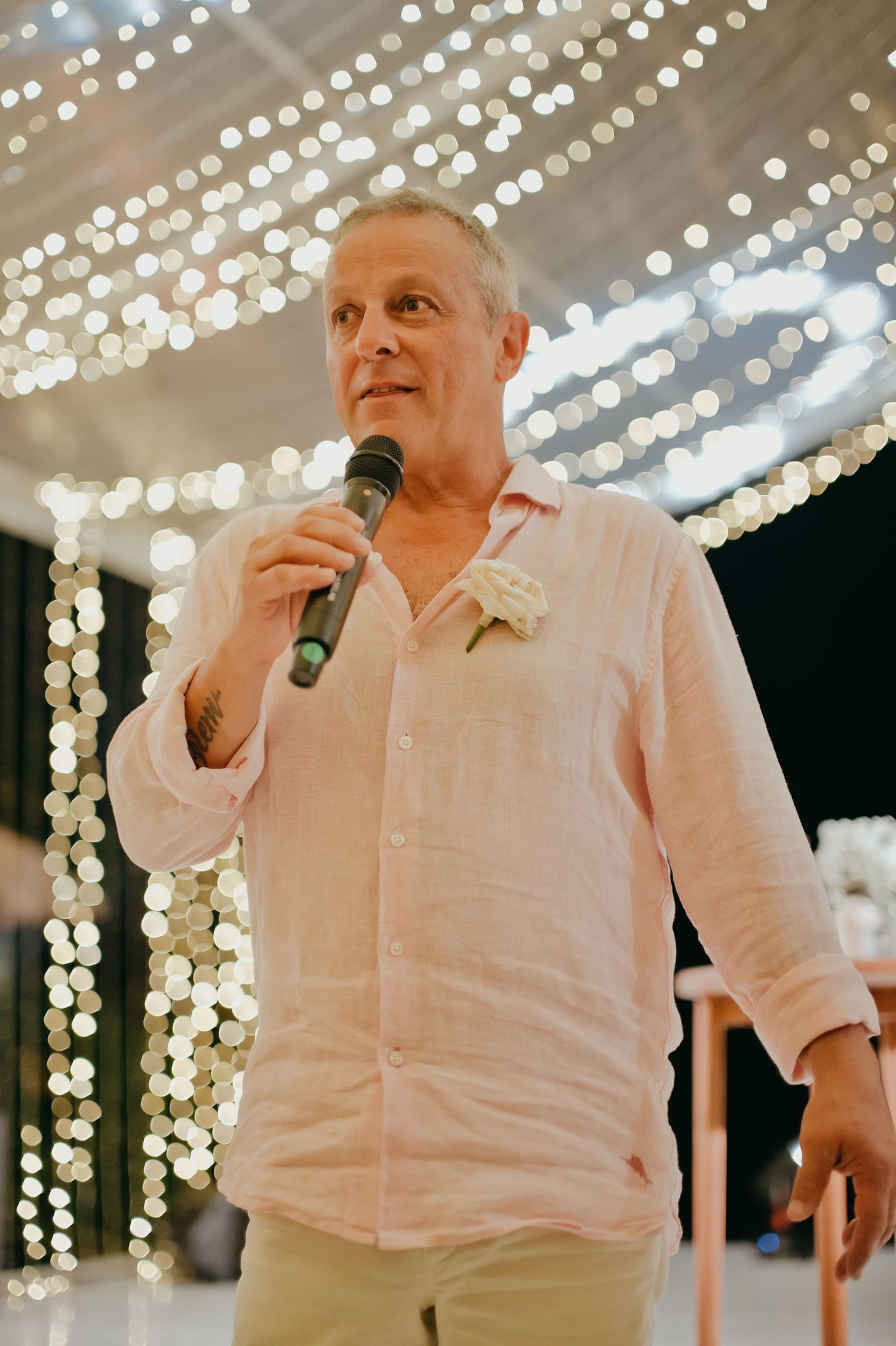 Man in pink linen shirt gives a heartfelt wedding speech under a canopy of warm fairy lights.