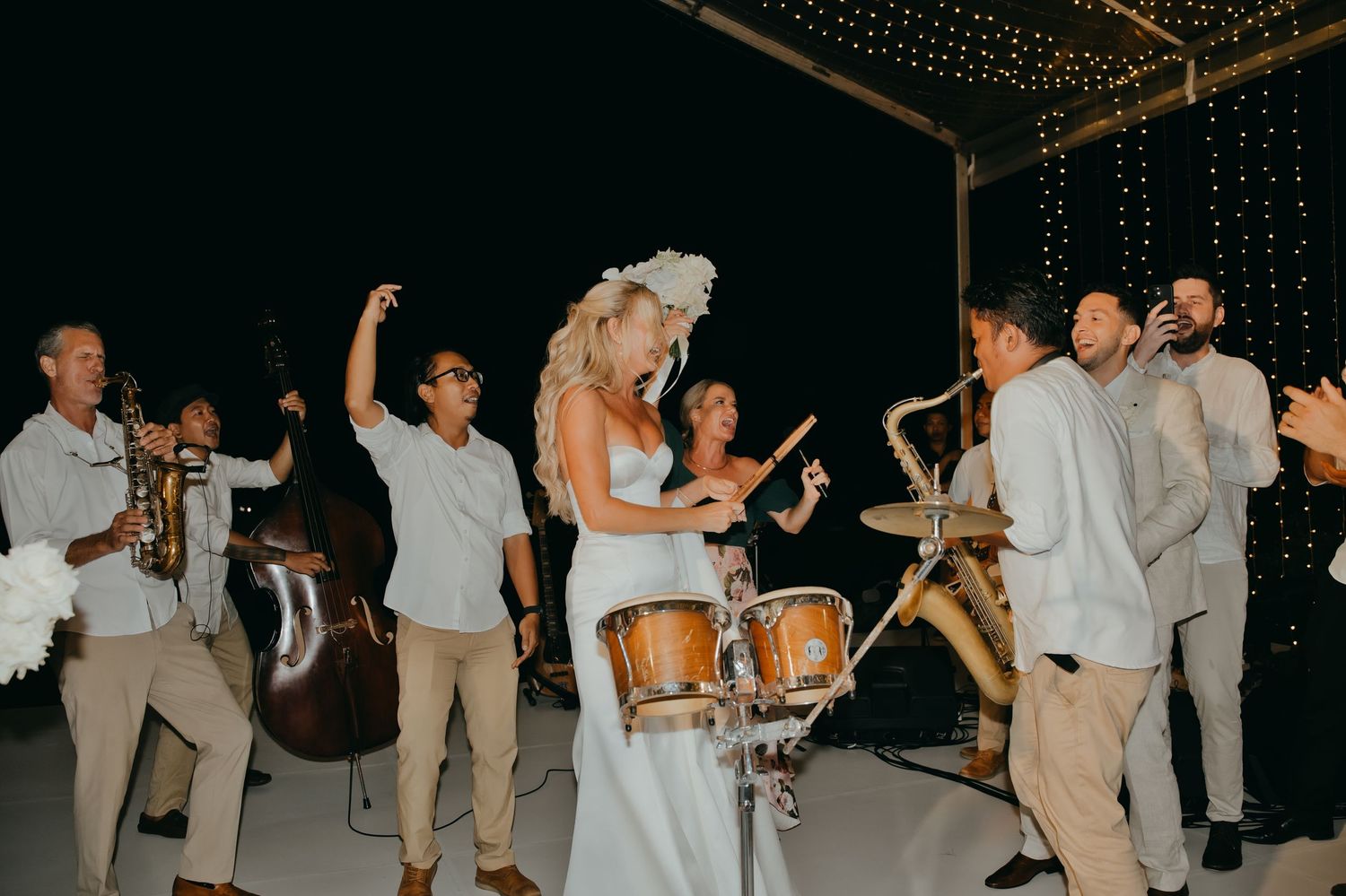 Bride dancing with live band musicians at an outdoor wedding reception under string lights.