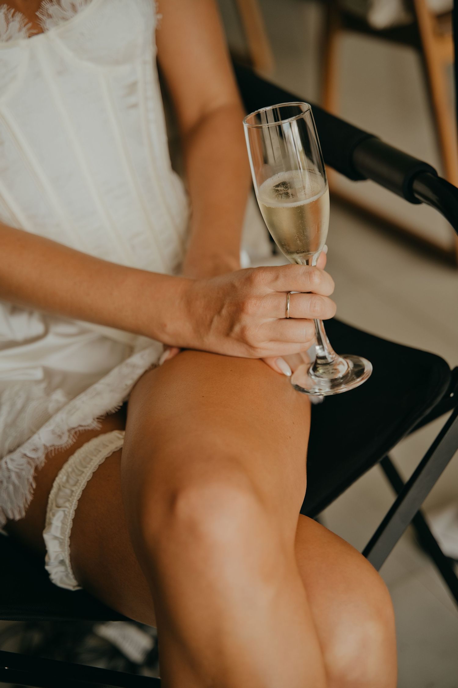 Bride in white lace dress holding champagne flute while seated, wearing wedding ring.