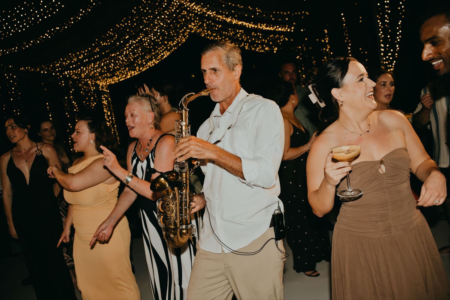 Saxophonist performs while guests dance joyfully under twinkling fairy lights at an outdoor wedding reception.