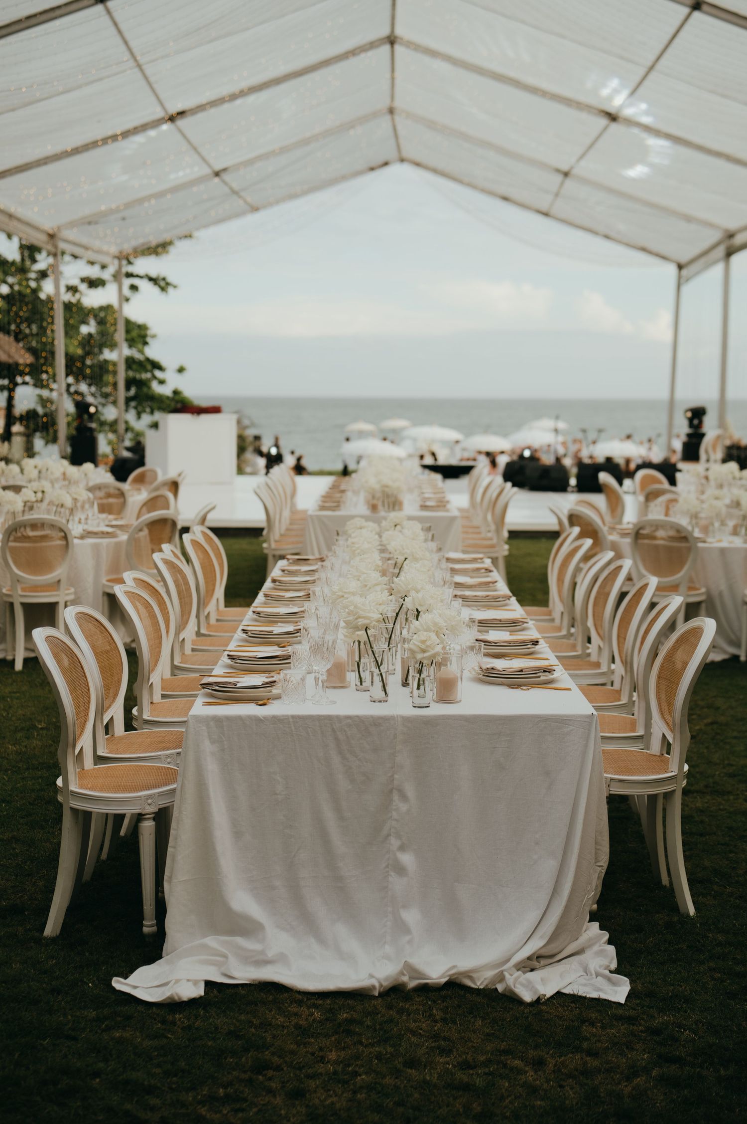 Elegant white reception tables set under a clear marquee tent with ocean views at sunset.