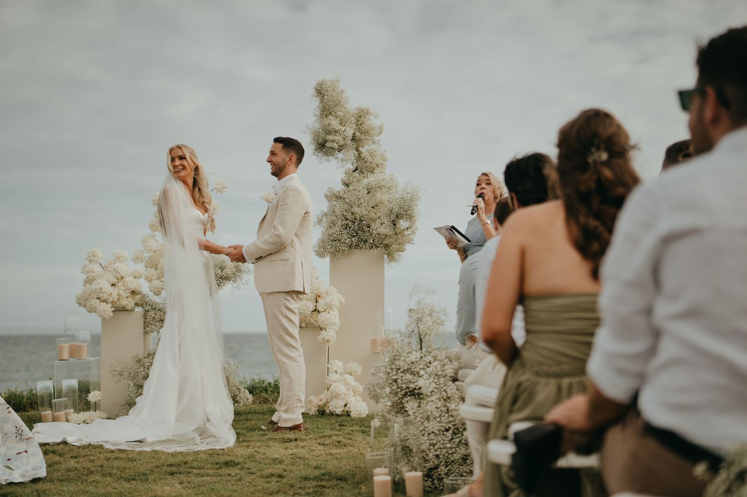 Bride and groom holding hands at outdoor oceanview wedding ceremony with white floral arrangements.