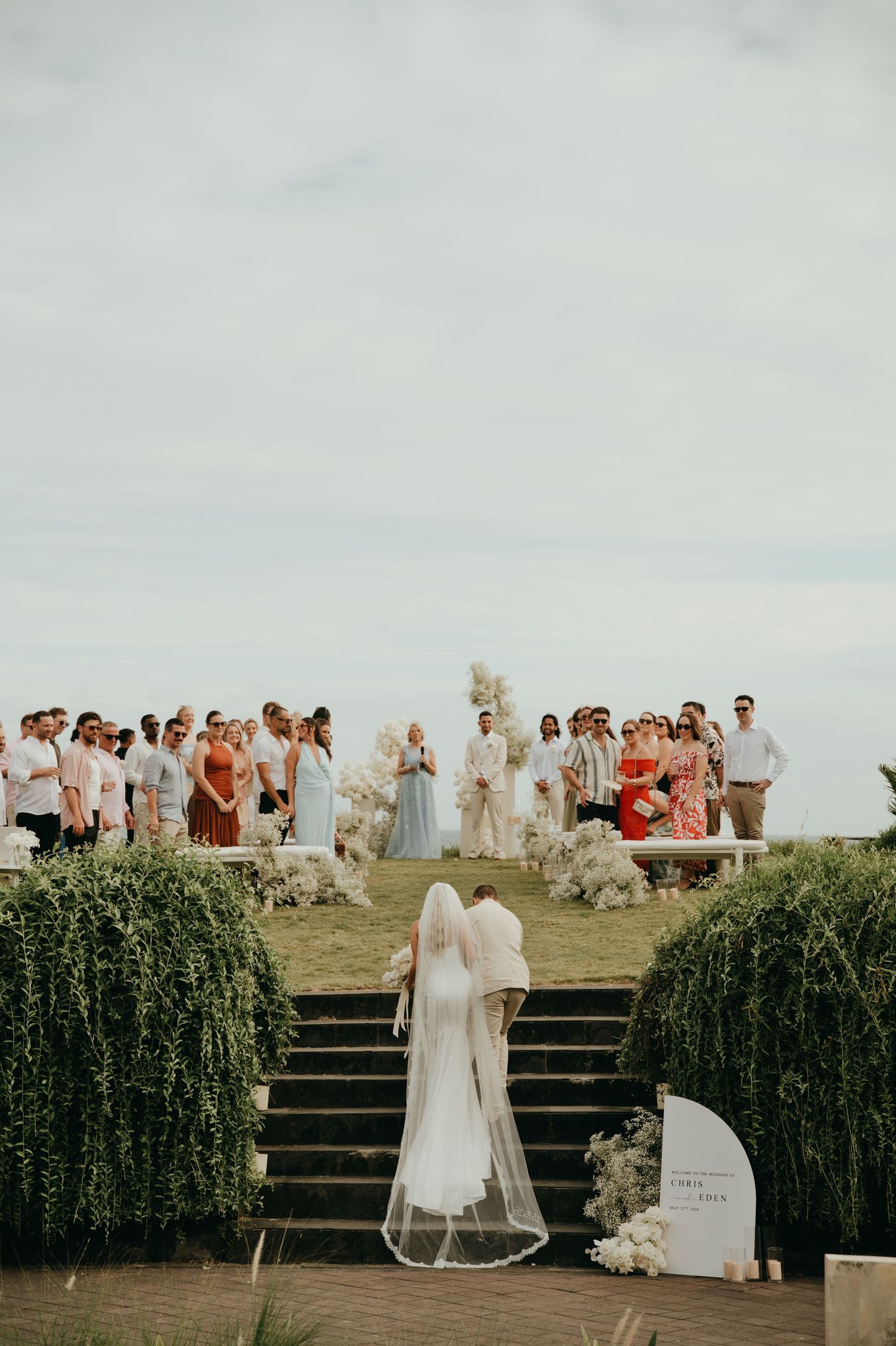 Bride in white gown and veil ascending garden stairs toward guests at outdoor clifftop ceremony.