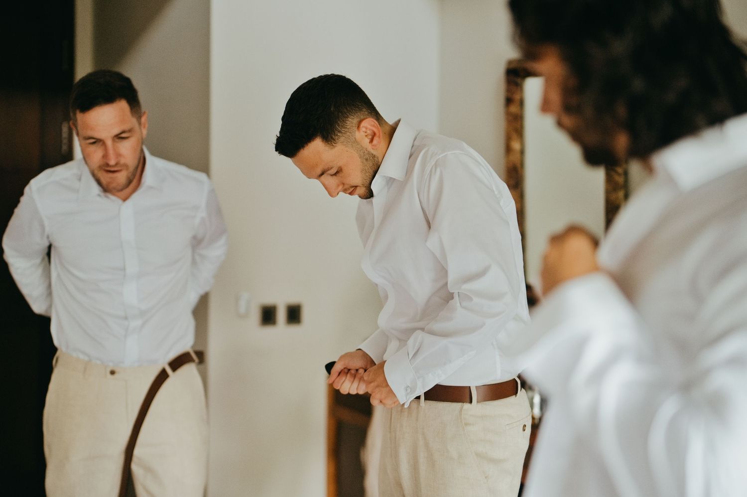 Groomsmen in white shirts and tan pants getting dressed together before wedding ceremony.
