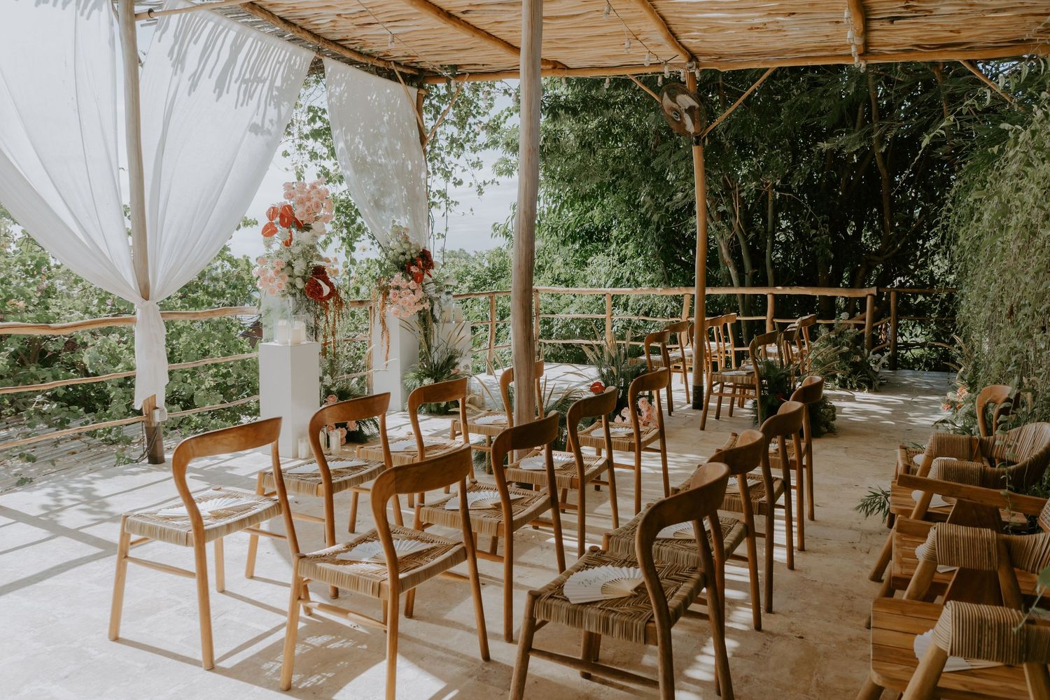 Outdoor wedding ceremony venue with wooden chairs, bamboo roof, white draping, and colorful floral altar arrangements.
