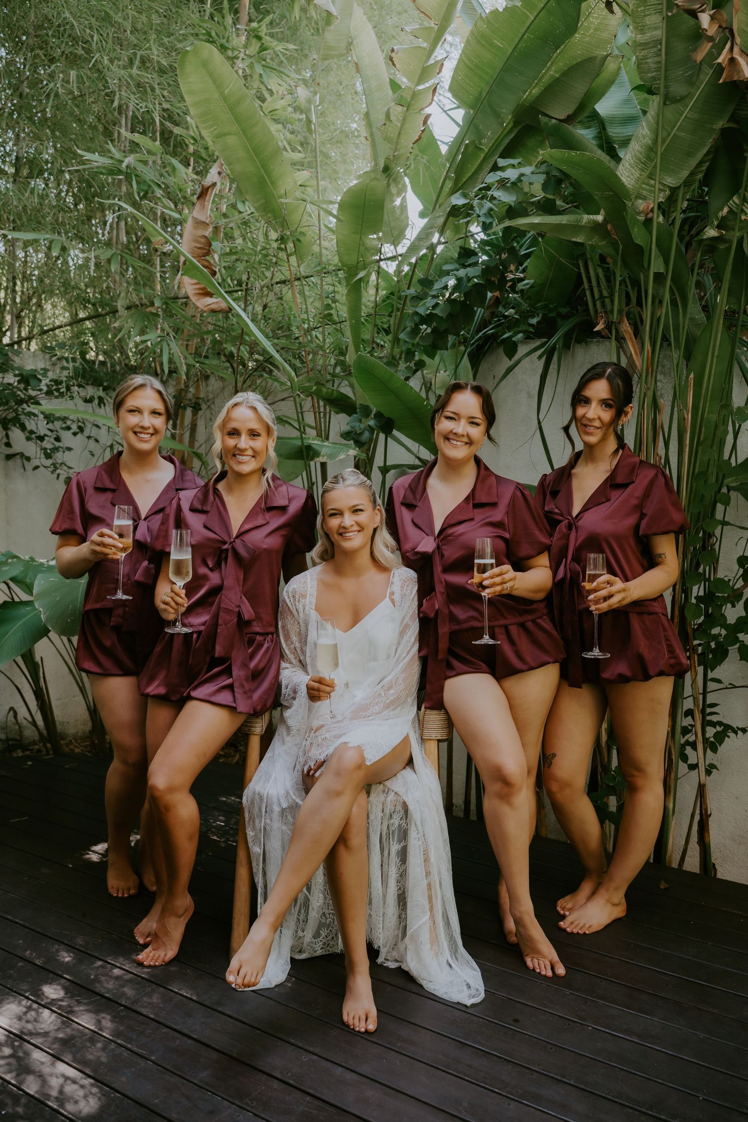 Bride in white robe sits with four bridesmaids in burgundy satin sets holding champagne glasses outdoors.