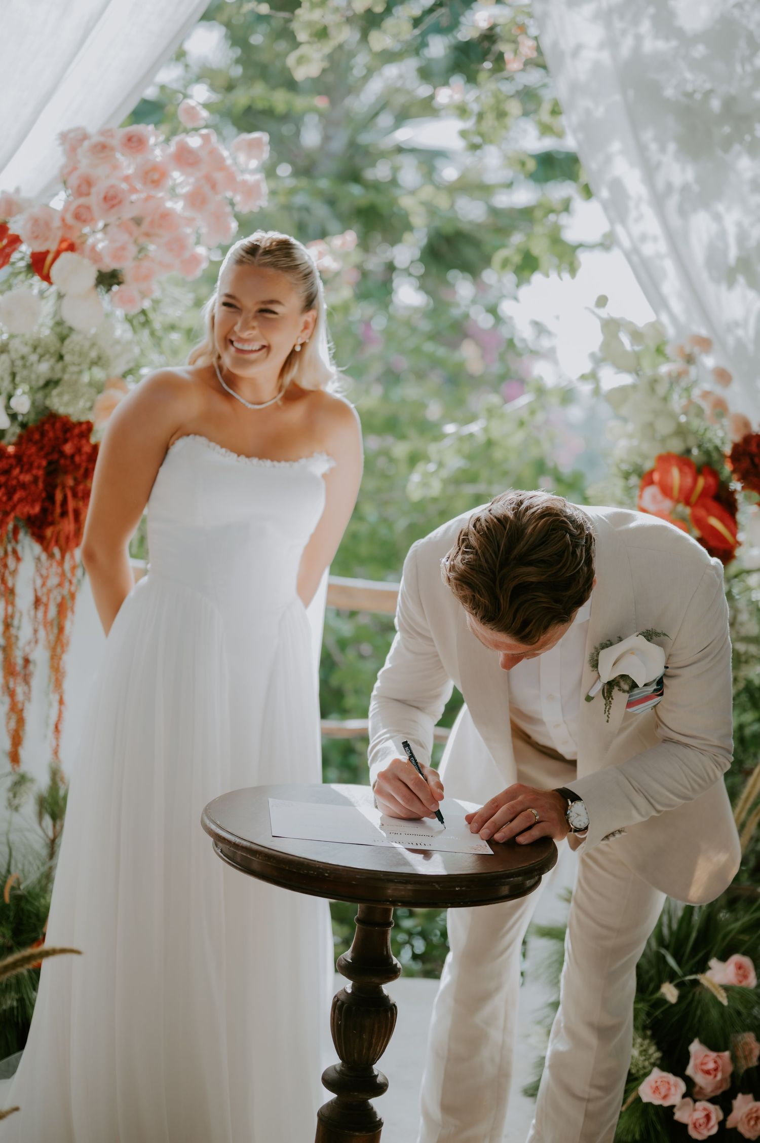 Groom signing wedding register as smiling bride in strapless gown watches at floral-decorated altar.