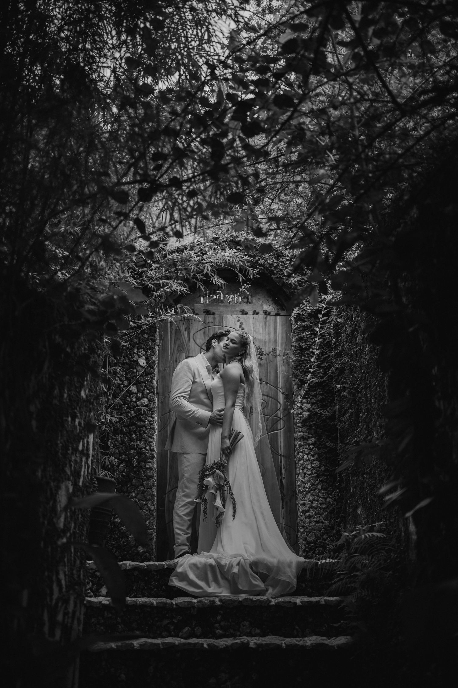Romantic black and white portrait of bride and groom kissing on ivy-covered stone steps.