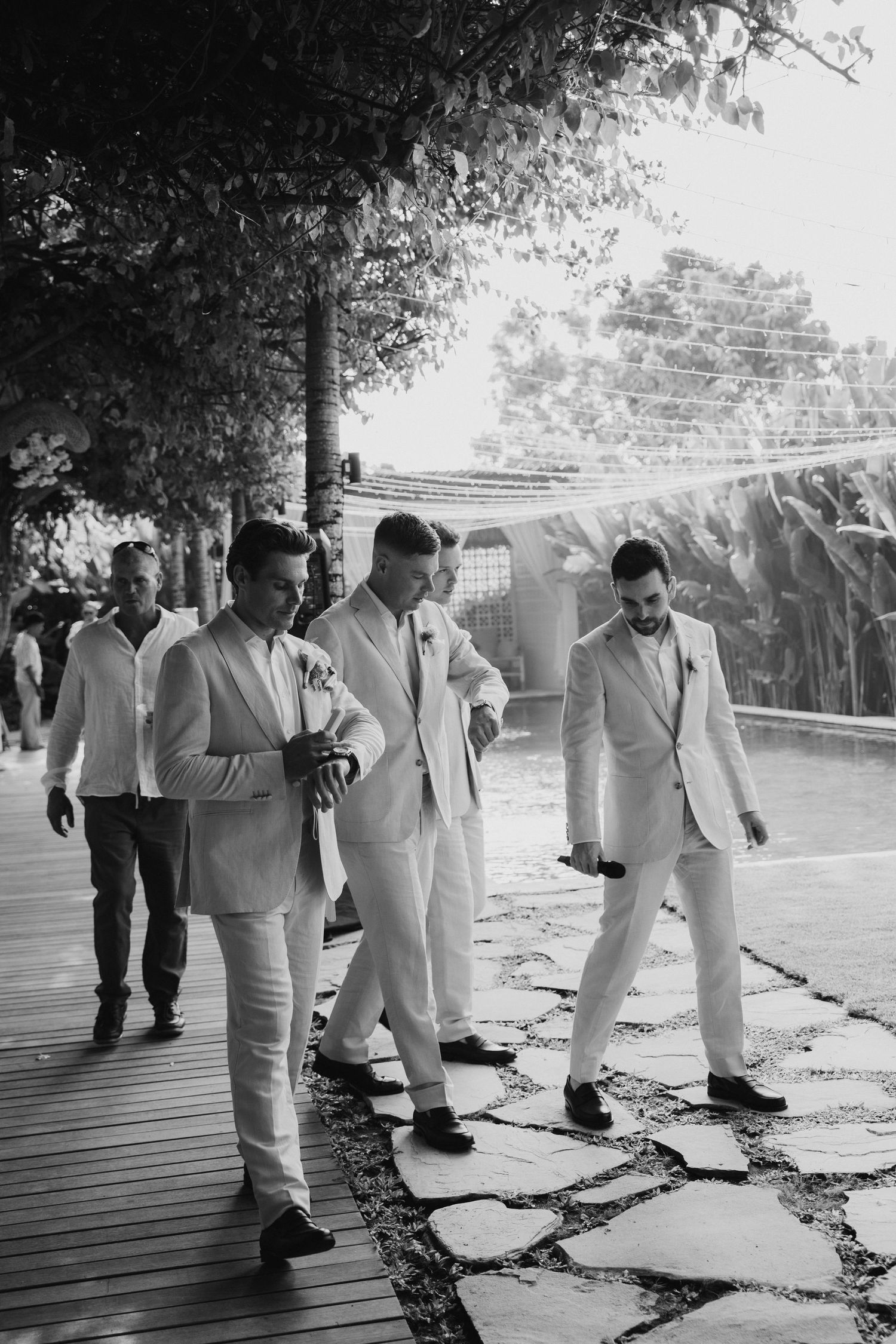 Groomsmen in white suits walking together on a stone pathway at an outdoor wedding venue.