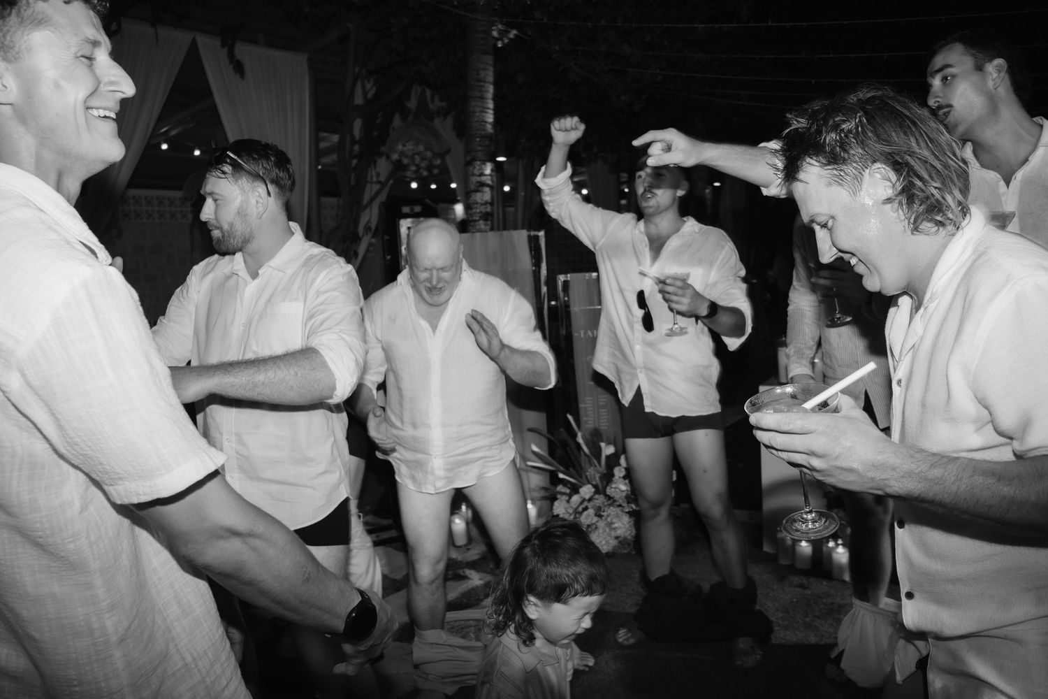 Black and white photo of wedding guests dancing and celebrating energetically at outdoor nighttime reception.