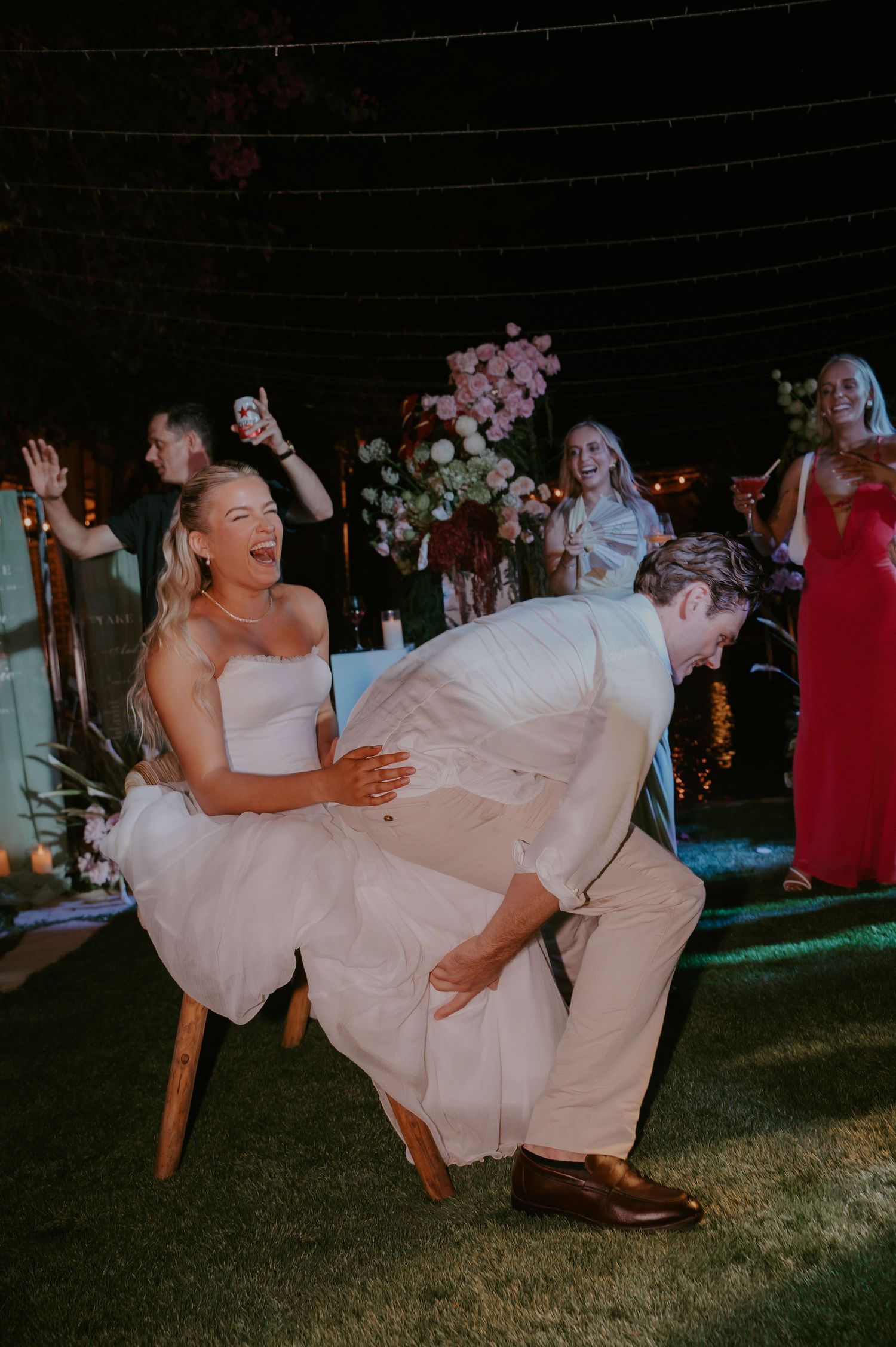 Laughing bride sits on chair while groom searches for garter during fun wedding reception tradition under string lights.