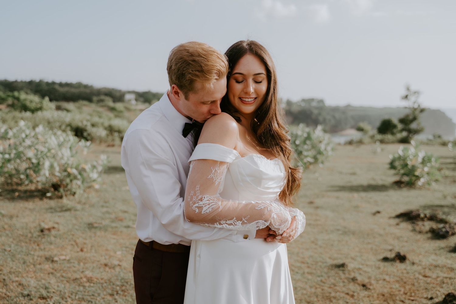 A groom in a white shirt embraces his bride in a white off-shoulder gown outdoors on their wedding day.