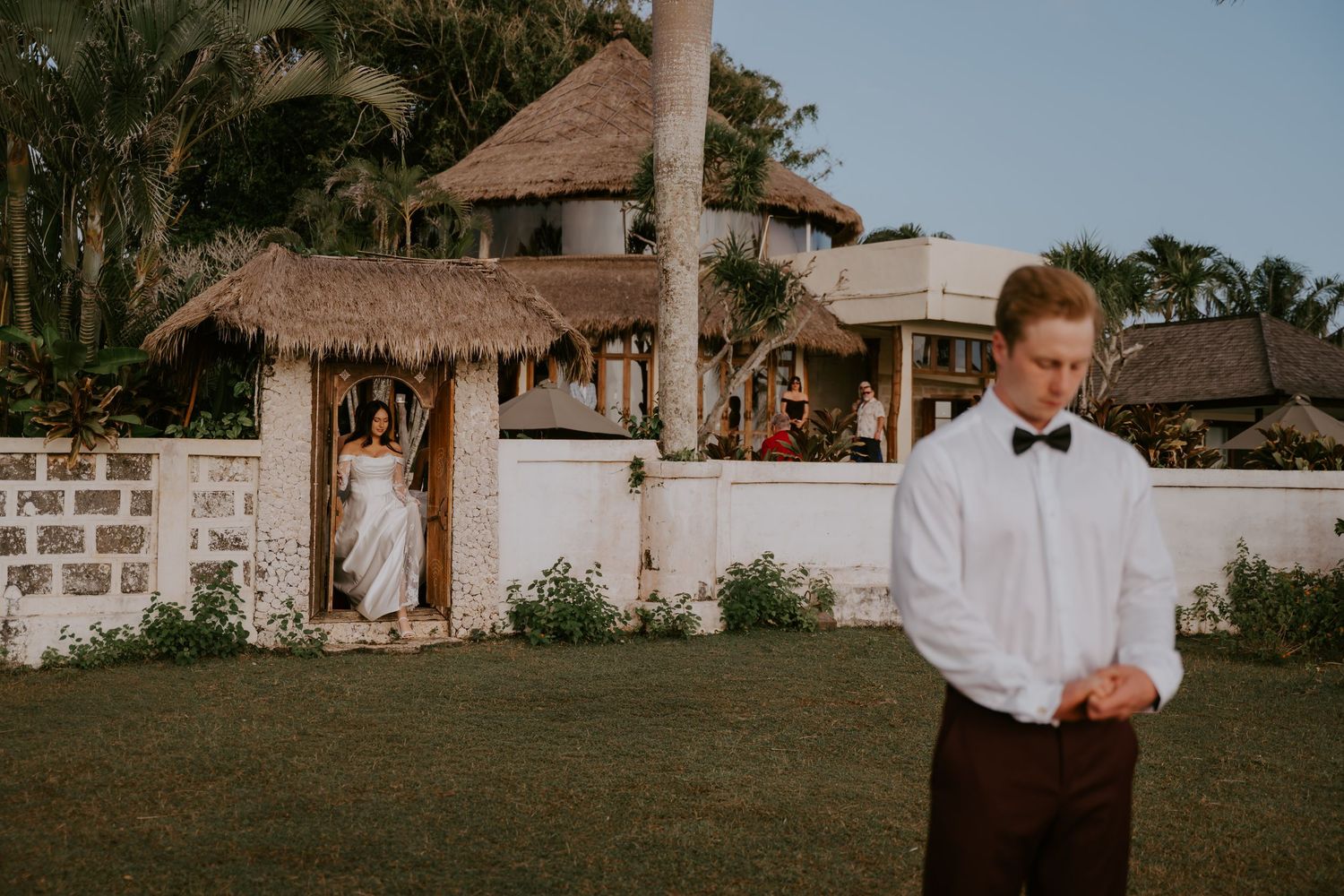 Groom in white shirt and bow tie stands on lawn as bride walks from thatched gazebo at tropical wedding venue.