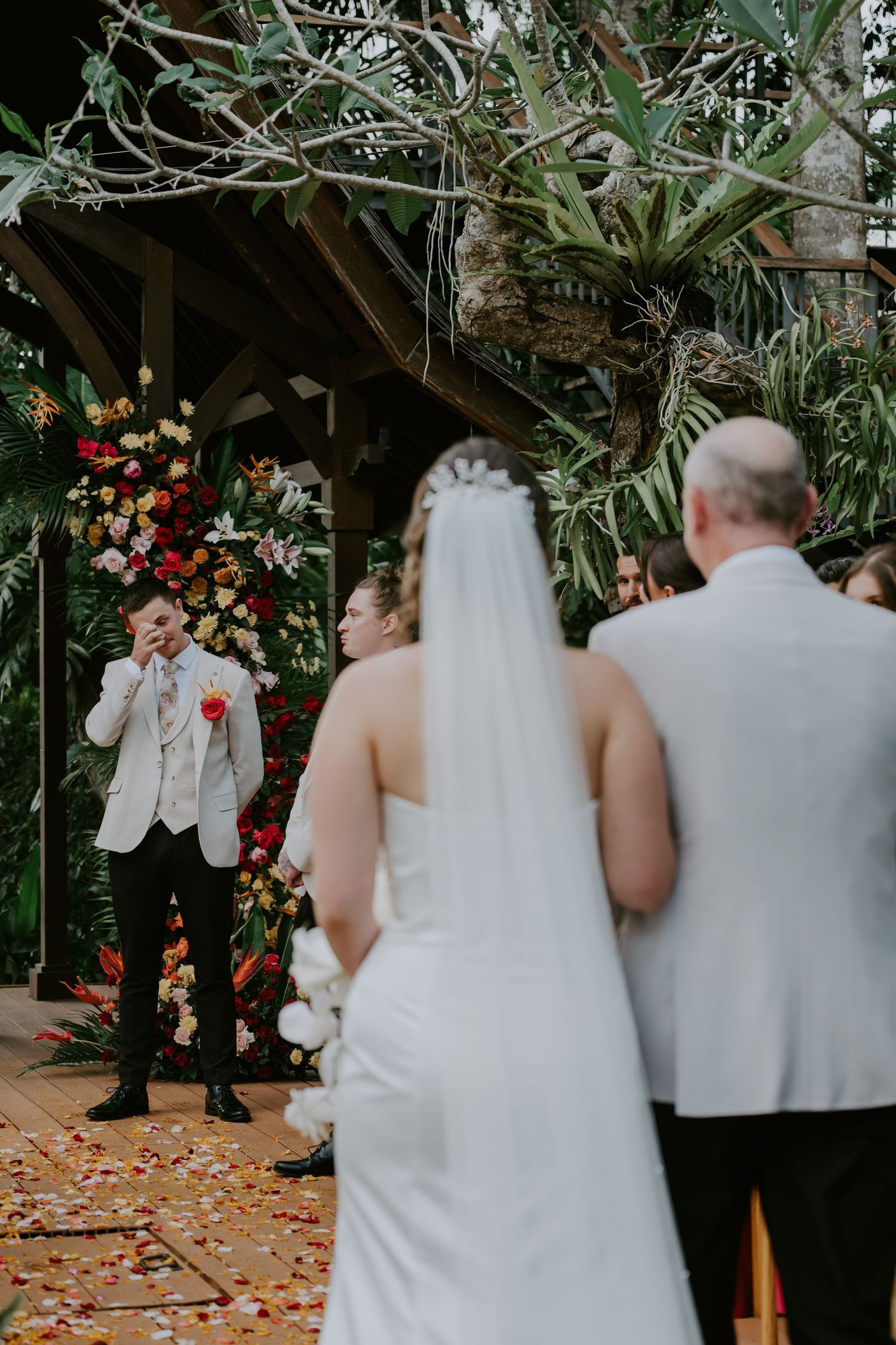 Emotional groom wipes tears watching bride walk down aisle toward vibrant tropical floral ceremony arch.