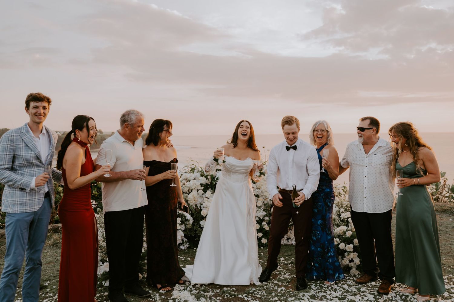Bride and groom laugh joyfully with family group portrait at stunning oceanside wedding at sunset.