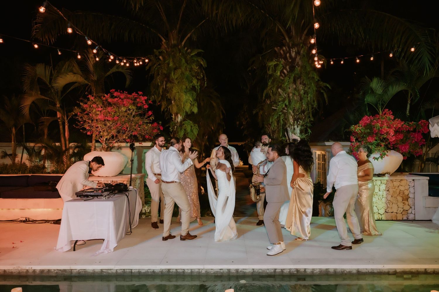 Wedding guests dance and celebrate poolside at night under string lights at tropical outdoor reception.