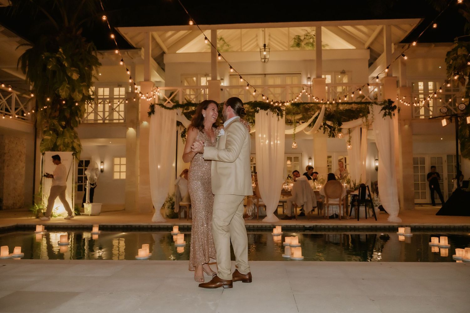 Bride and groom share a romantic first dance beside a pool with floating candles at night.