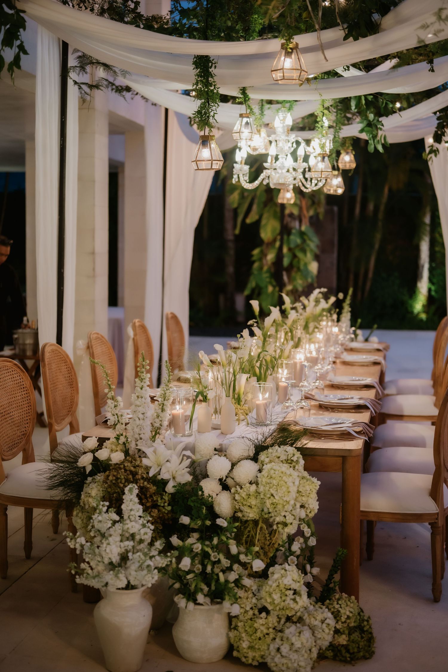 Elegant wedding reception table adorned with white flowers, candles, and crystal chandeliers at night.