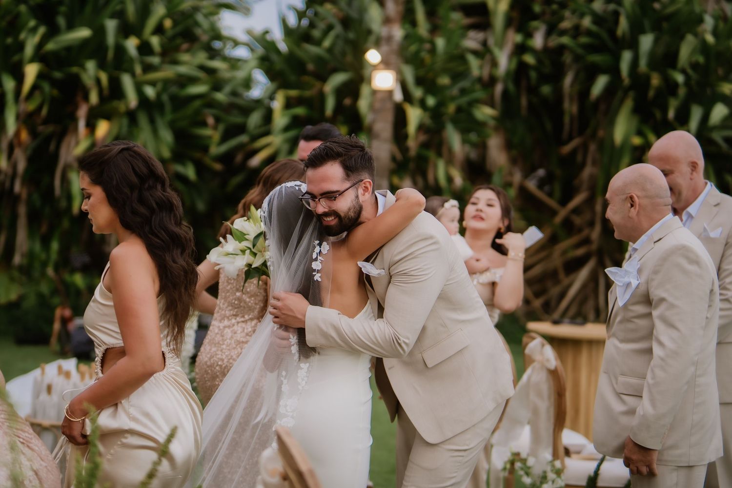 The groom embraces a guest warmly while the bride stands nearby after the ceremony.